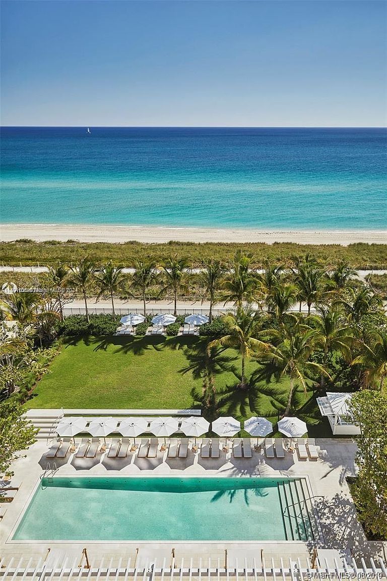 This is an aerial view showcasing a luxurious pool area with a pristine turquoise pool, surrounded by lounge chairs and white umbrellas. Beyond the pool, a manicured green lawn leads to palm trees and a sandy beach with the ocean in the background. The scene evokes a sense of relaxation and upscale coastal living.