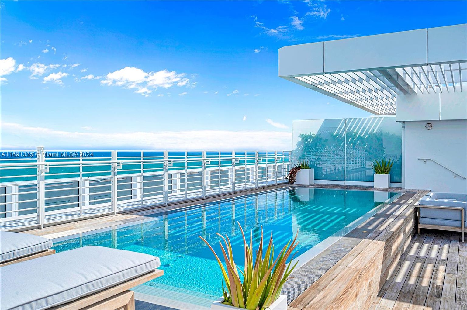 This image showcases a luxurious rooftop pool area with stunning ocean views. The pool is surrounded by modern architectural elements, including a pergola and glass railings. Lounge chairs and a seating area provide ample space for relaxation, creating an inviting and upscale atmosphere.