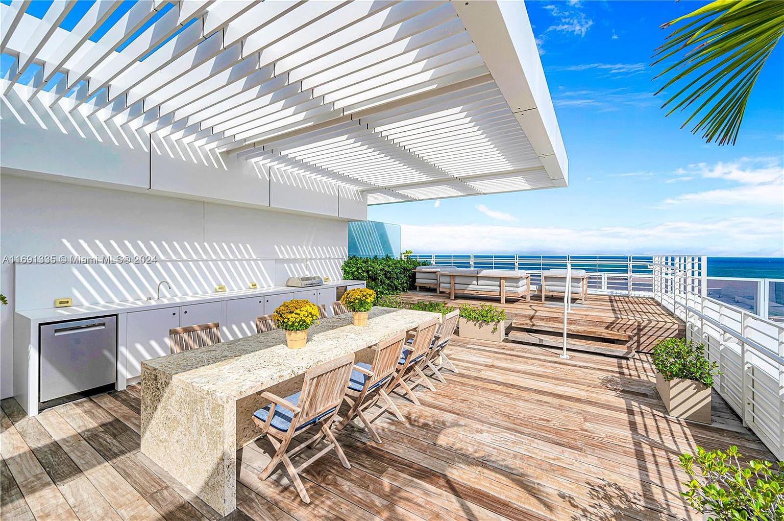 This is a luxurious outdoor patio/deck/balcony area with a stunning ocean view. The space features a long dining table with seating, an outdoor kitchen area, and a lounge area with comfortable seating. The wooden deck and white pergola create a bright and inviting atmosphere, perfect for entertaining or relaxing.