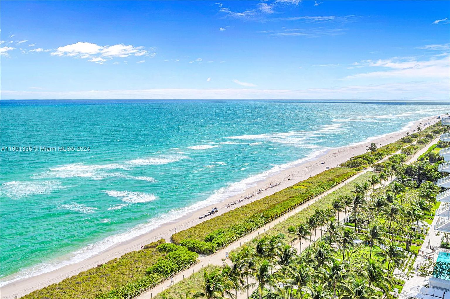 This aerial view showcases a stunning coastline with turquoise waters meeting a pristine sandy beach. Lush green vegetation lines the shore, complemented by palm trees and glimpses of residential buildings. The scene evokes a sense of luxury and relaxation, highlighting the desirable beachfront location.