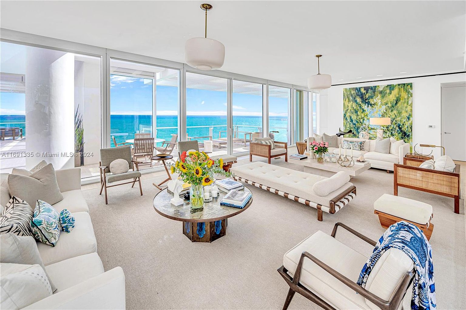 This is a bright and airy living room with floor-to-ceiling windows offering stunning ocean views. The room is furnished with comfortable, modern furniture in neutral tones, including sofas, chairs, and a daybed. A large abstract painting adds a touch of color to the space, and the overall impression is one of luxury and relaxation.