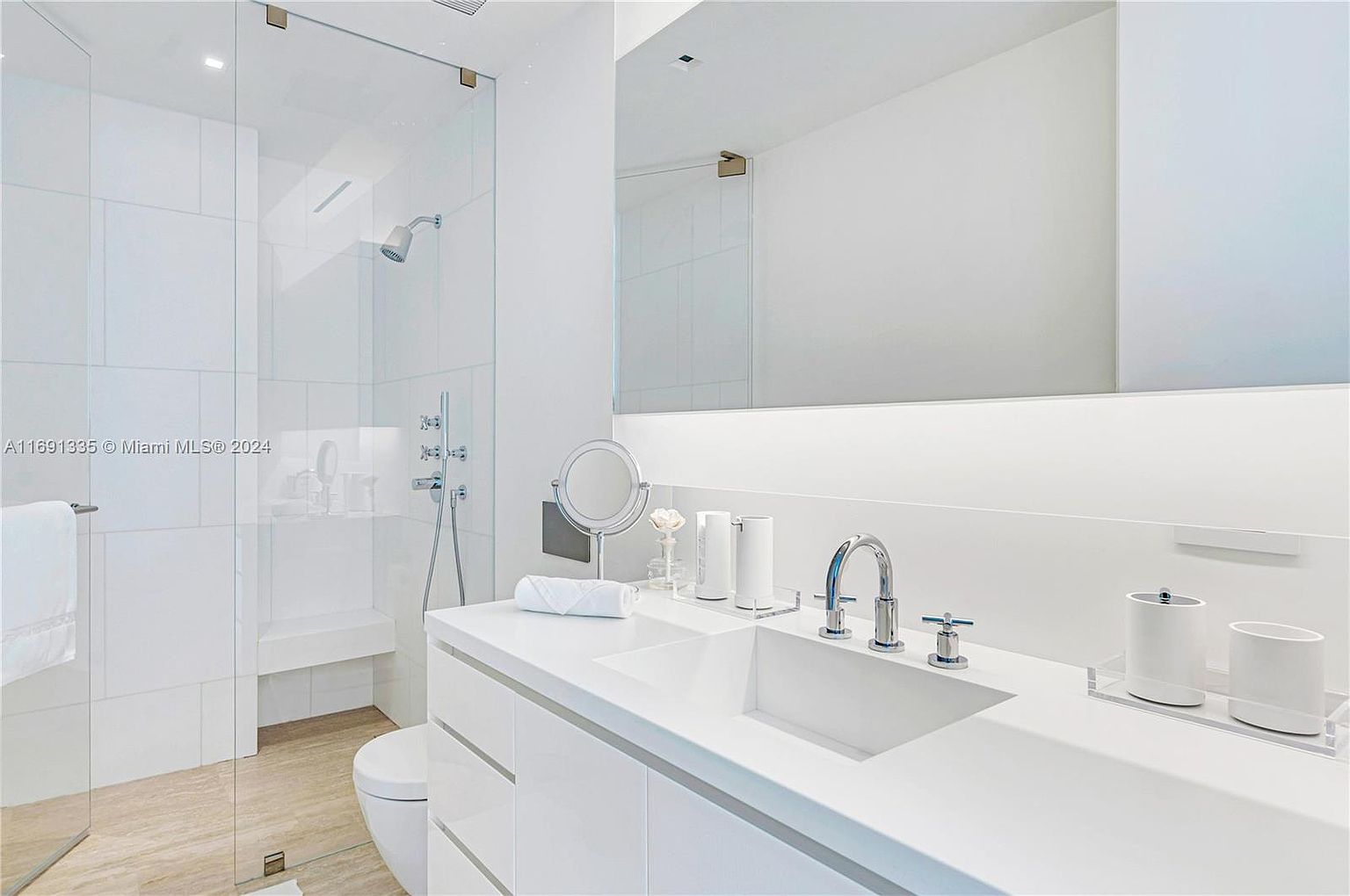 This is a bright and modern primary bathroom featuring a glass-enclosed shower with a built-in bench, a sleek white vanity with dual sinks, and a large mirror. The space is illuminated by integrated lighting and natural light, creating a clean and luxurious atmosphere. The perspective is from the doorway, showcasing the entire bathroom.