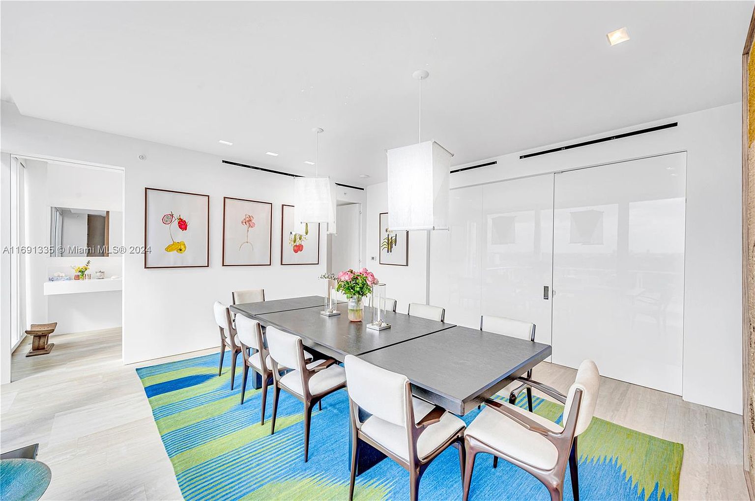This is a bright and modern dining room featuring a long, dark gray dining table with seating for ten. The room is decorated with framed artwork and illuminated by contemporary pendant lights. A vibrant blue and green rug adds a pop of color to the light wood flooring, creating an inviting and stylish space.