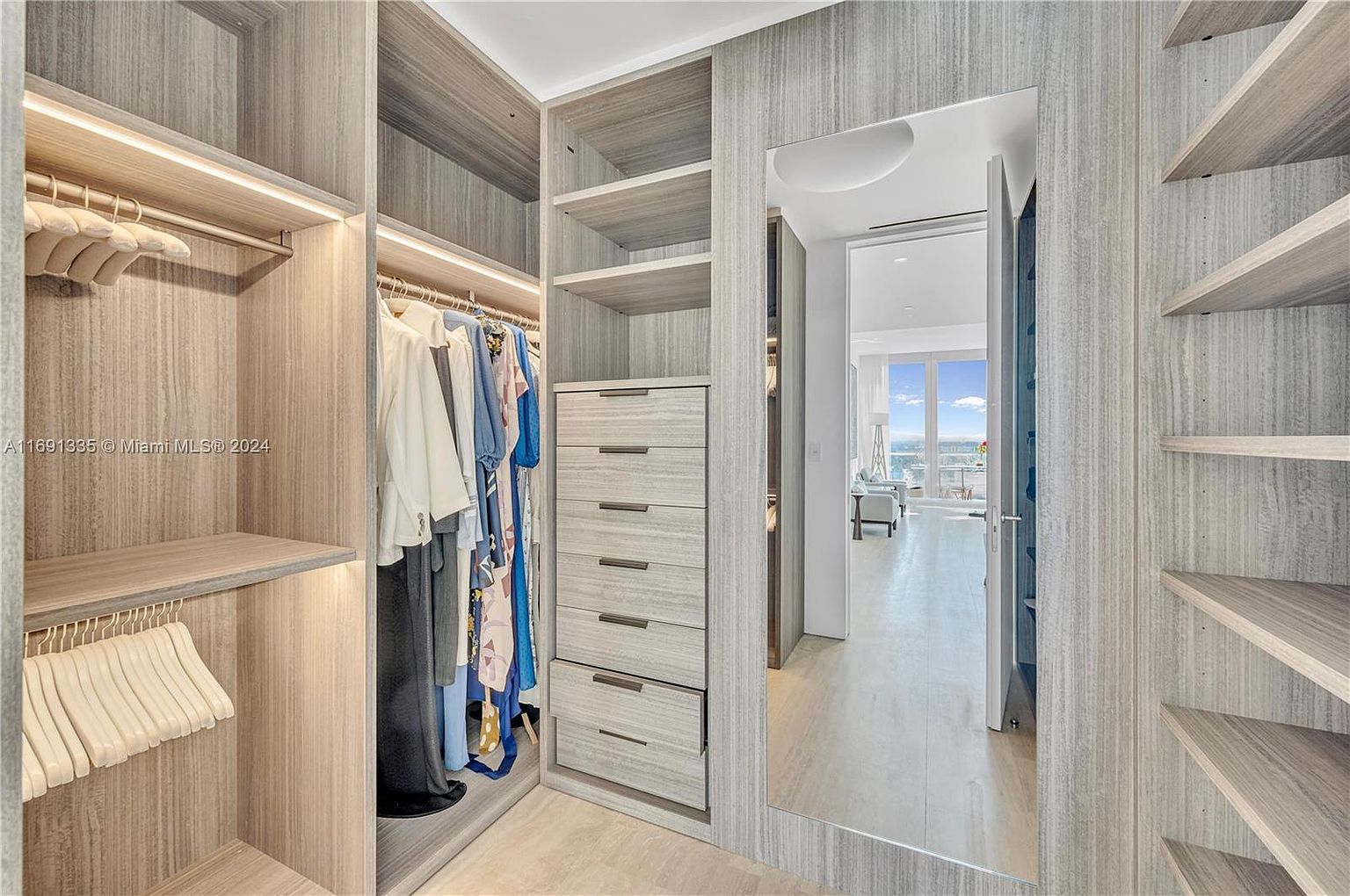 This image showcases a well-organized walk-in closet with custom shelving, drawers, and hanging rods. The closet features a neutral color palette with wood-grain finishes, creating a sophisticated and functional space. A full-length mirror reflects the adjacent living area, enhancing the sense of openness and luxury.