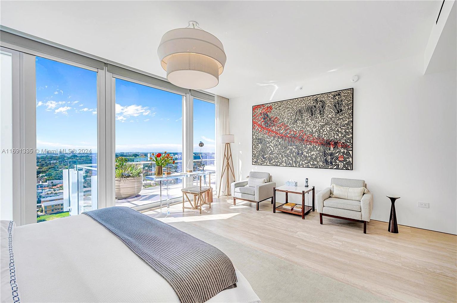 This is a bright and airy primary bedroom with floor-to-ceiling windows offering a stunning view. The room features a large bed with a gray throw, two armchairs with a small table between them, and a striking piece of artwork on the wall. The light wood flooring and neutral color palette create a serene and luxurious atmosphere.