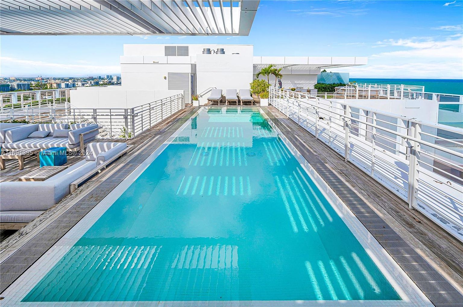 This image showcases a luxurious rooftop pool area, featuring a long, rectangular pool with clear blue water reflecting the sky and surrounding architecture. Modern white lounge furniture is arranged on the wooden deck, complemented by sleek white railings and a pergola-style roof structure. The scene offers a sense of upscale relaxation and stunning city and ocean views.