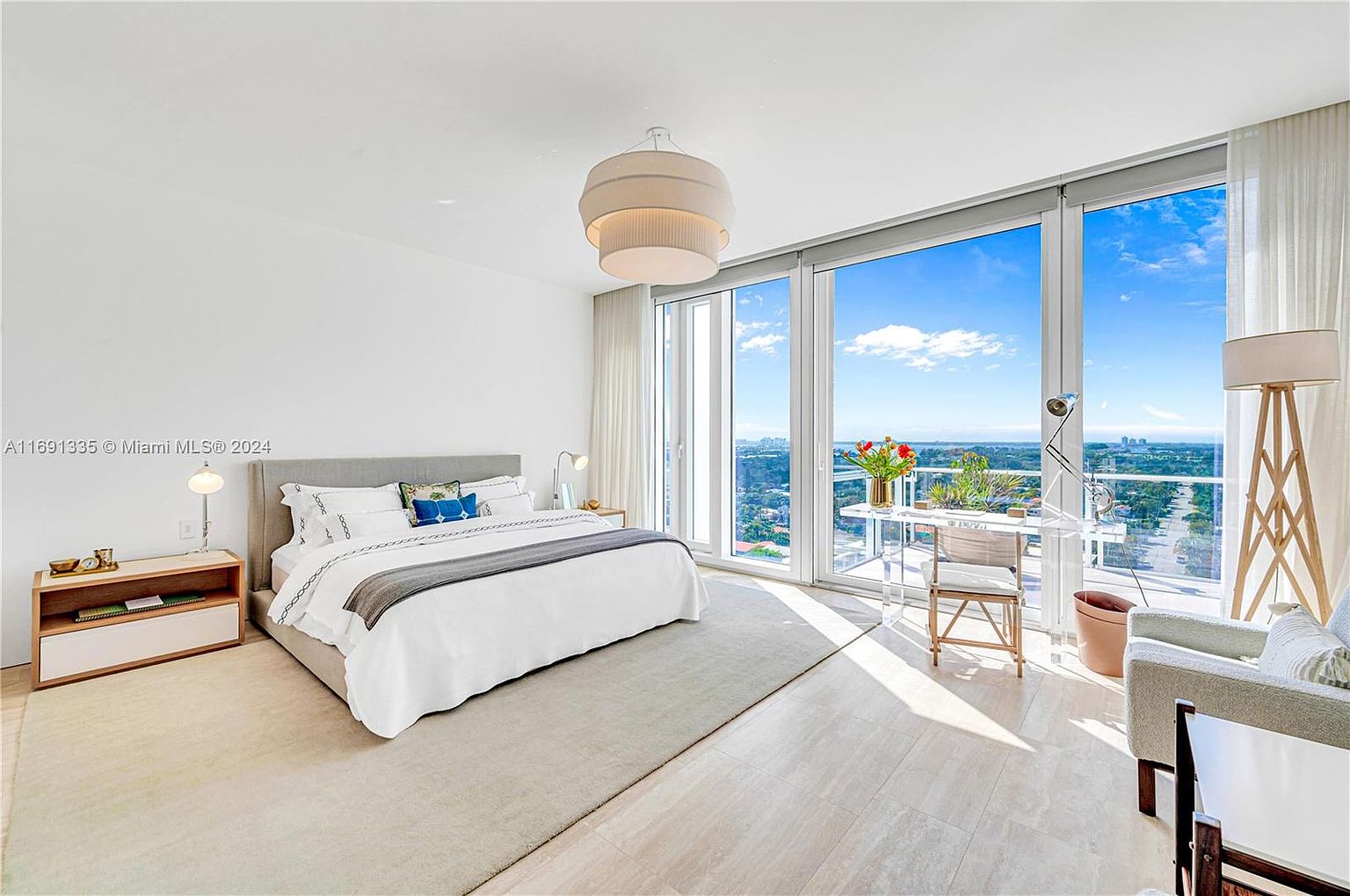 This is a bright and airy primary bedroom featuring a large bed with neutral linens, a modern bedside table, and a floor lamp. The room boasts floor-to-ceiling windows offering stunning views of the city and sky, complemented by sheer curtains. A clear desk and chair are positioned near the windows, creating a workspace with a view.