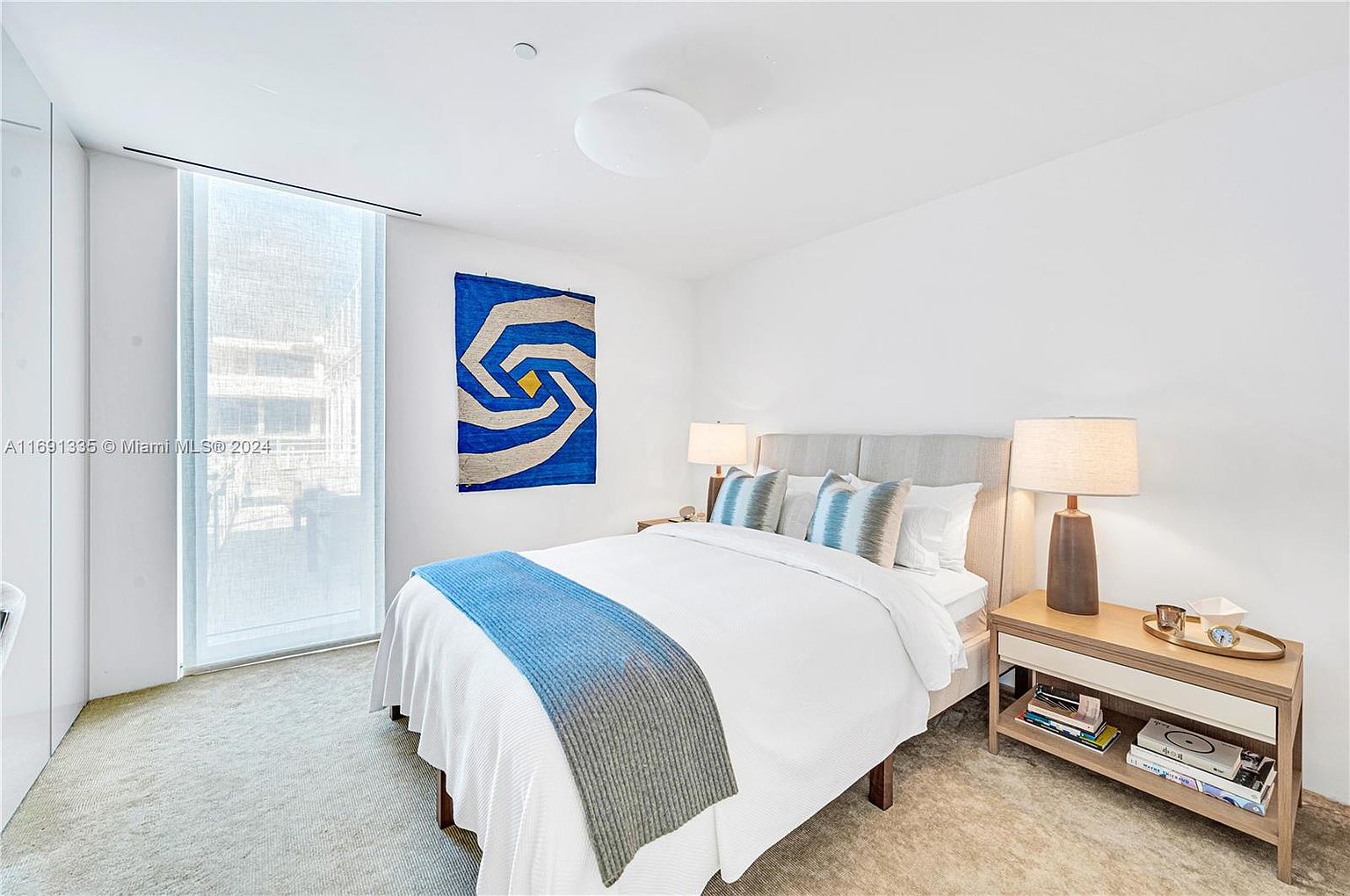 This is a bright and airy primary bedroom featuring a queen-sized bed with white linens, a blue and gray throw, and decorative pillows. A modern art piece hangs above the bed, and a light wood nightstand with a lamp sits to the right. Natural light floods the room through a large window, creating a serene and inviting atmosphere.