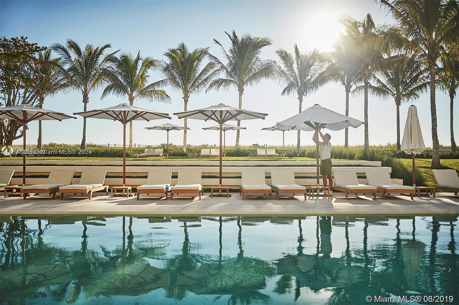 The image showcases a luxurious pool area with multiple lounge chairs and umbrellas, set against a backdrop of palm trees and a clear sky. The pool's water reflects the surrounding scenery, creating a serene and inviting atmosphere. This outdoor space is designed for relaxation and leisure, highlighting the property's upscale amenities.