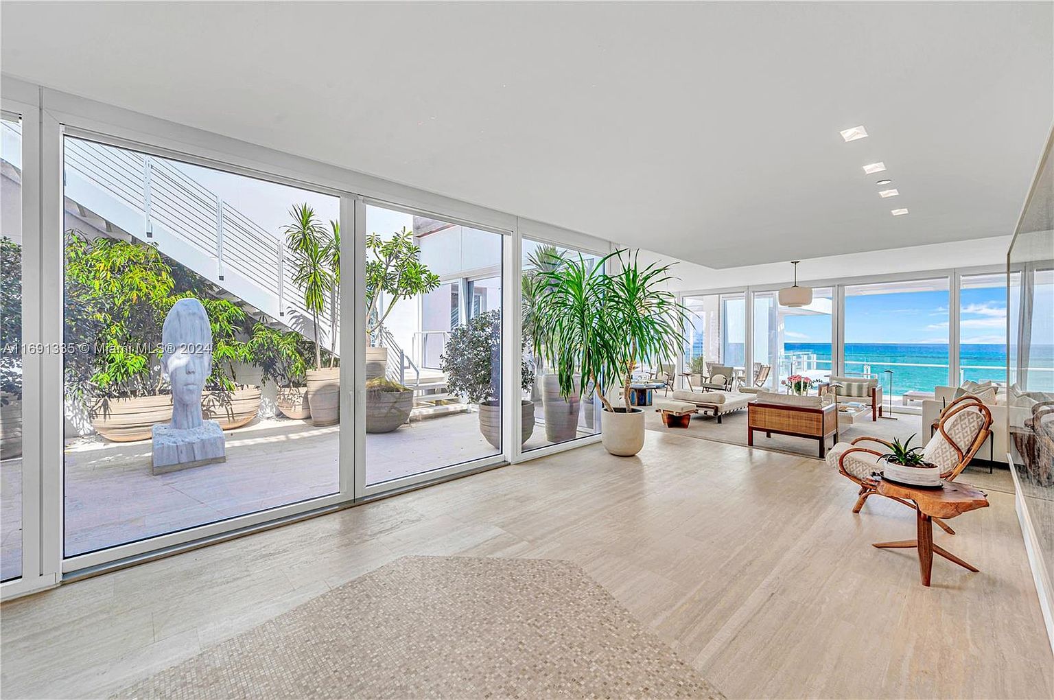 This is a bright and airy living room with floor-to-ceiling windows offering stunning ocean views. The room features a mix of modern and natural elements, including a unique wooden chair, neutral-toned furniture, and large potted plants that bring the outdoors in. The open floor plan and abundant natural light create a luxurious and inviting atmosphere.