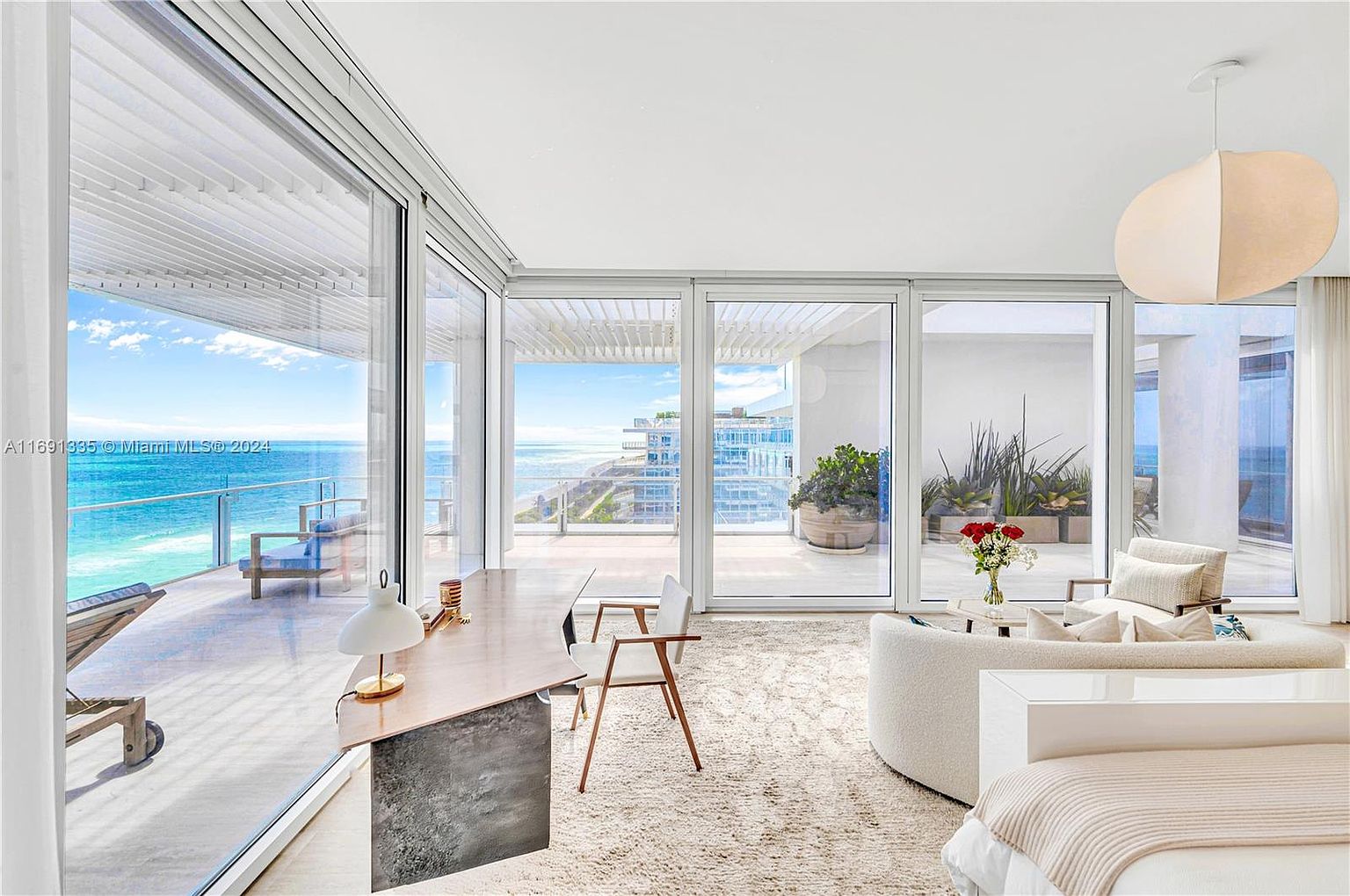 This is a bright and airy living room with floor-to-ceiling windows offering stunning ocean views. The room features a modern aesthetic with a neutral color palette, a curved sofa, and a unique desk. The space is designed to maximize natural light and showcase the beautiful coastal scenery.