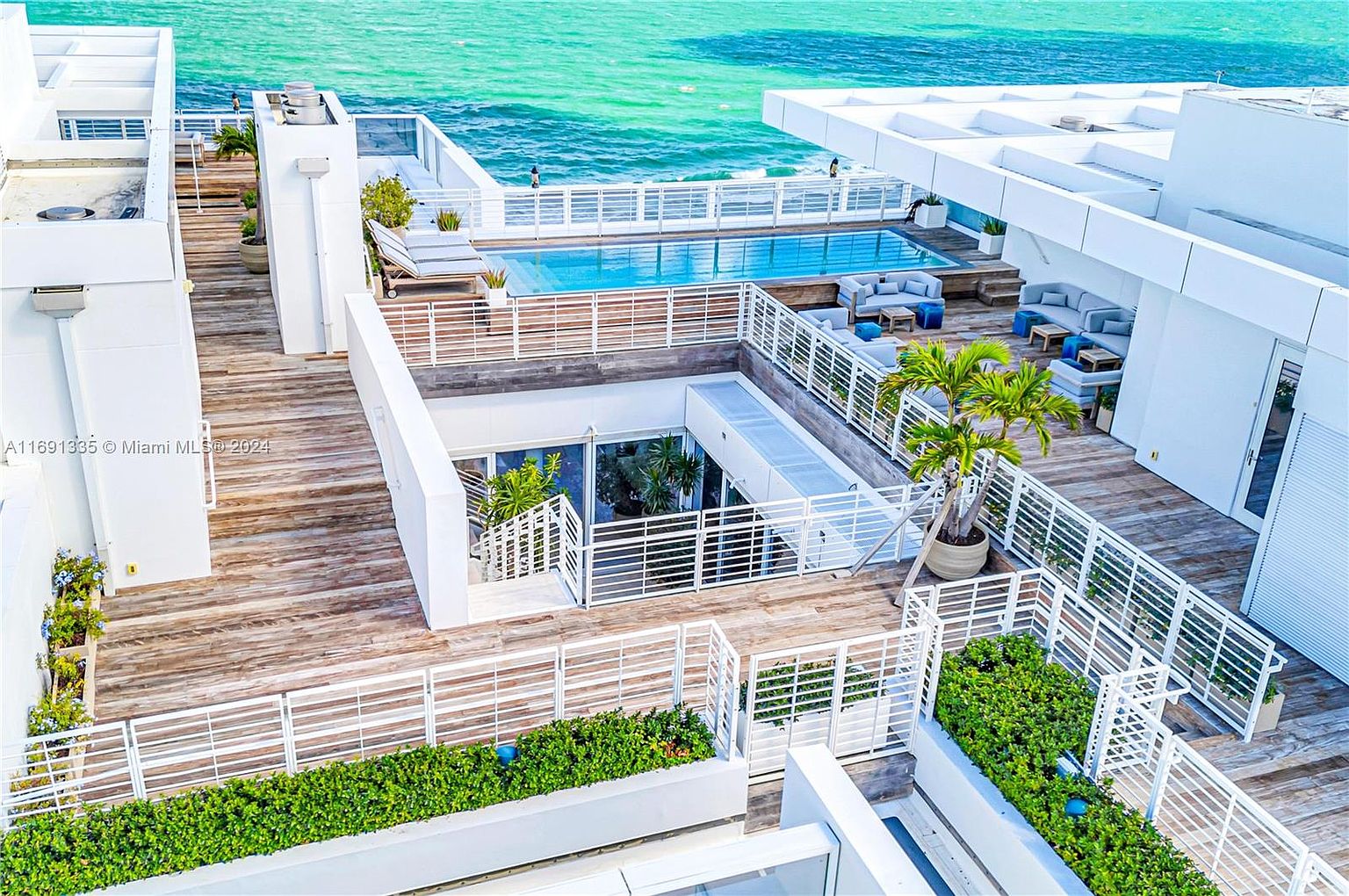 This aerial view showcases a luxurious rooftop deck with a swimming pool overlooking the ocean. The deck features multiple seating areas with modern outdoor furniture, lush greenery, and white architectural elements, creating a sophisticated and relaxing atmosphere. The overall impression is one of high-end coastal living.