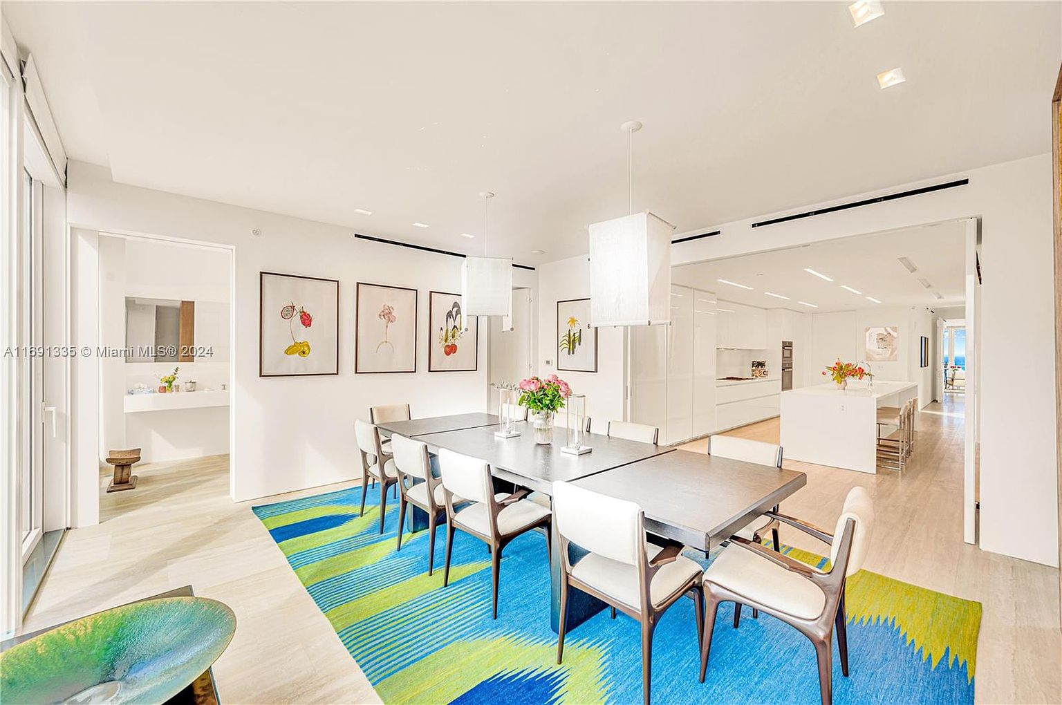 This is an interior shot of a modern dining room. The room features a long dining table with chairs, a colorful rug, and artwork on the walls. The space is well-lit with pendant lighting and natural light, creating a bright and inviting atmosphere, with a view into the kitchen.