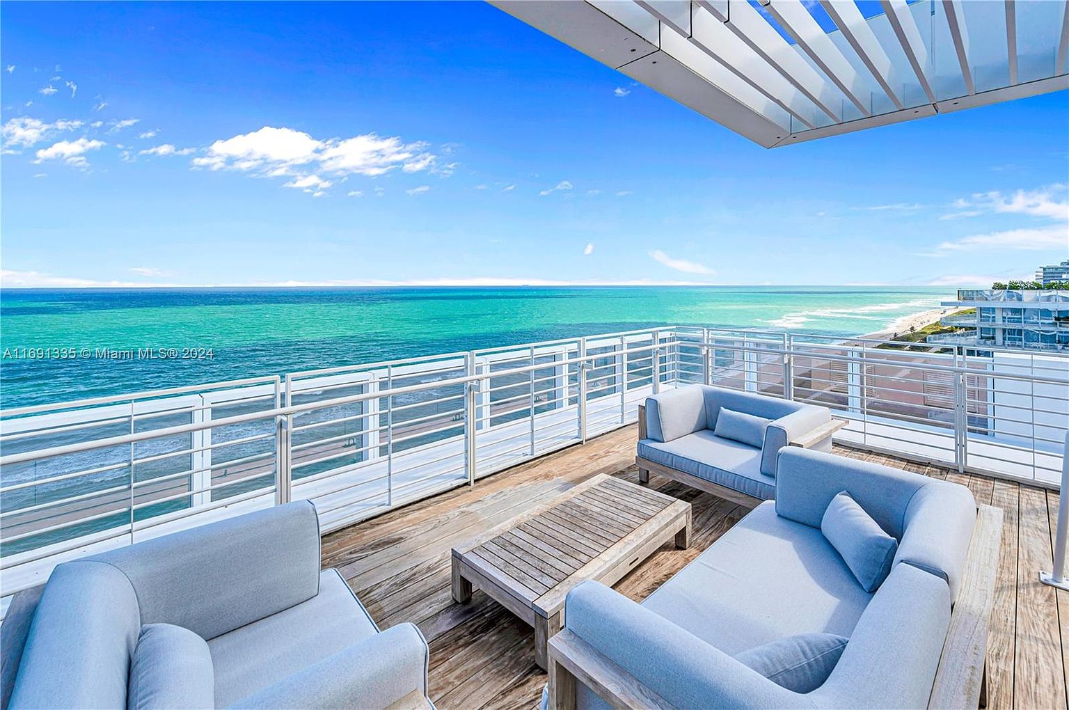 This image showcases a luxurious outdoor living space on a balcony or deck overlooking the ocean. The area is furnished with modern, light-colored sofas and a wooden coffee table, creating a comfortable and stylish lounge area. A white railing provides safety and unobstructed views of the turquoise water and sandy beach, enhancing the property's appeal for relaxation and entertainment.