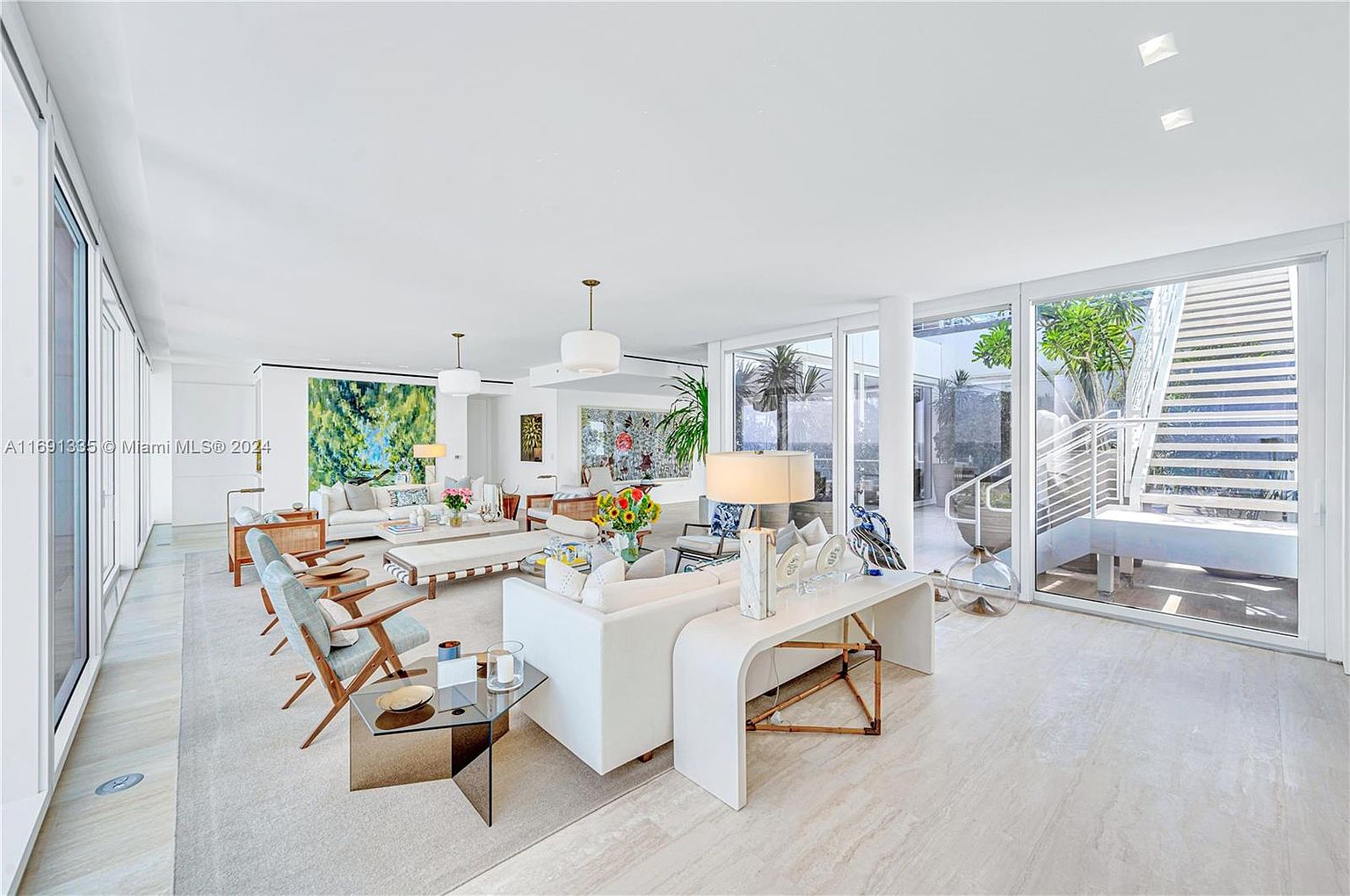 This is a bright and spacious living room featuring a modern design with white walls and light-colored flooring. The room is furnished with multiple white sofas, wooden chairs, and glass tables, creating a comfortable and inviting atmosphere. Large windows and sliding glass doors provide ample natural light and access to an outdoor area with stairs.