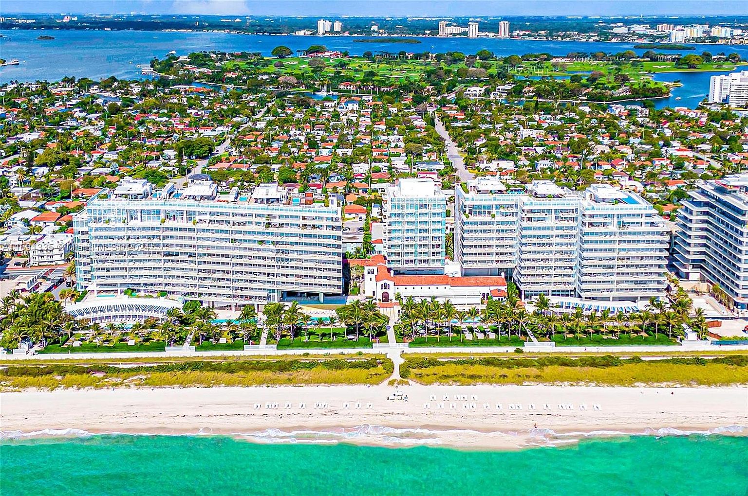 This aerial view showcases a luxurious beachfront property featuring multiple modern, white condominium buildings. The buildings are situated along a pristine beach with turquoise waters, complemented by lush landscaping and palm trees. The overall impression is one of high-end coastal living with stunning ocean views and meticulously maintained grounds.