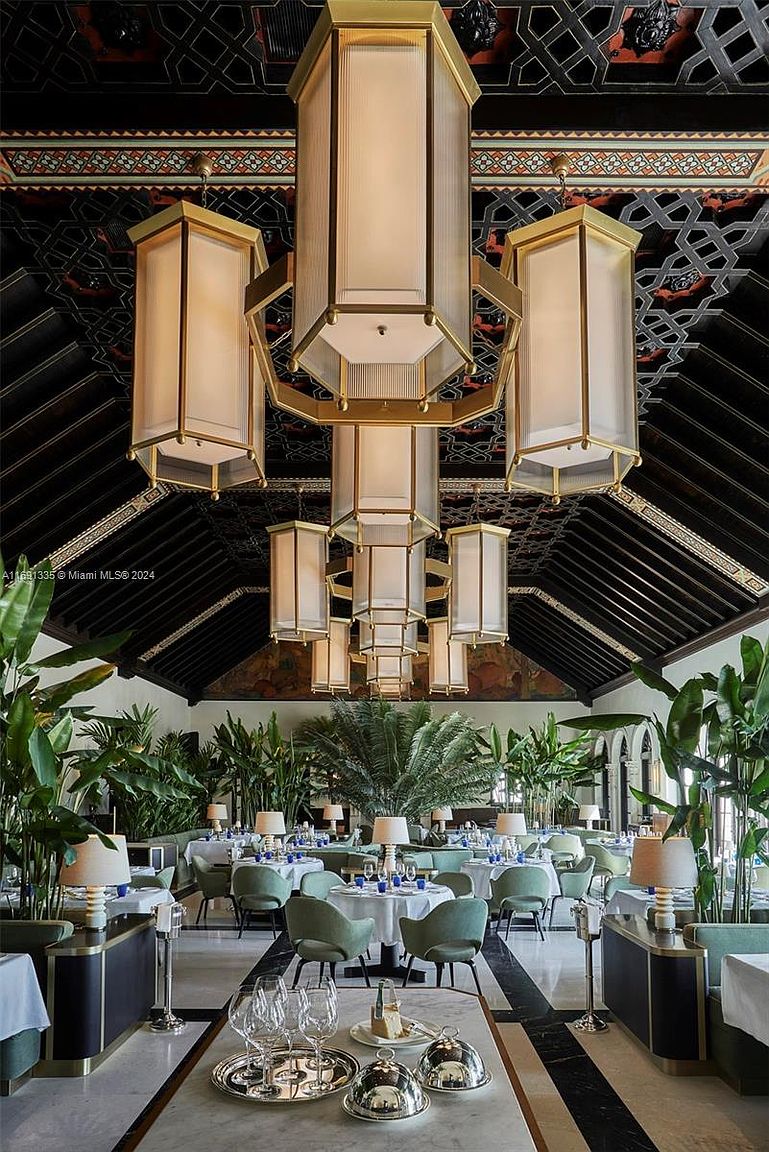 This is an interior shot of an elegant dining room featuring a high, intricately decorated ceiling and large, ornate chandeliers. The room is filled with round tables set for dining, surrounded by comfortable chairs and lush greenery. The overall impression is one of luxury and sophistication, perfect for a high-end dining experience.