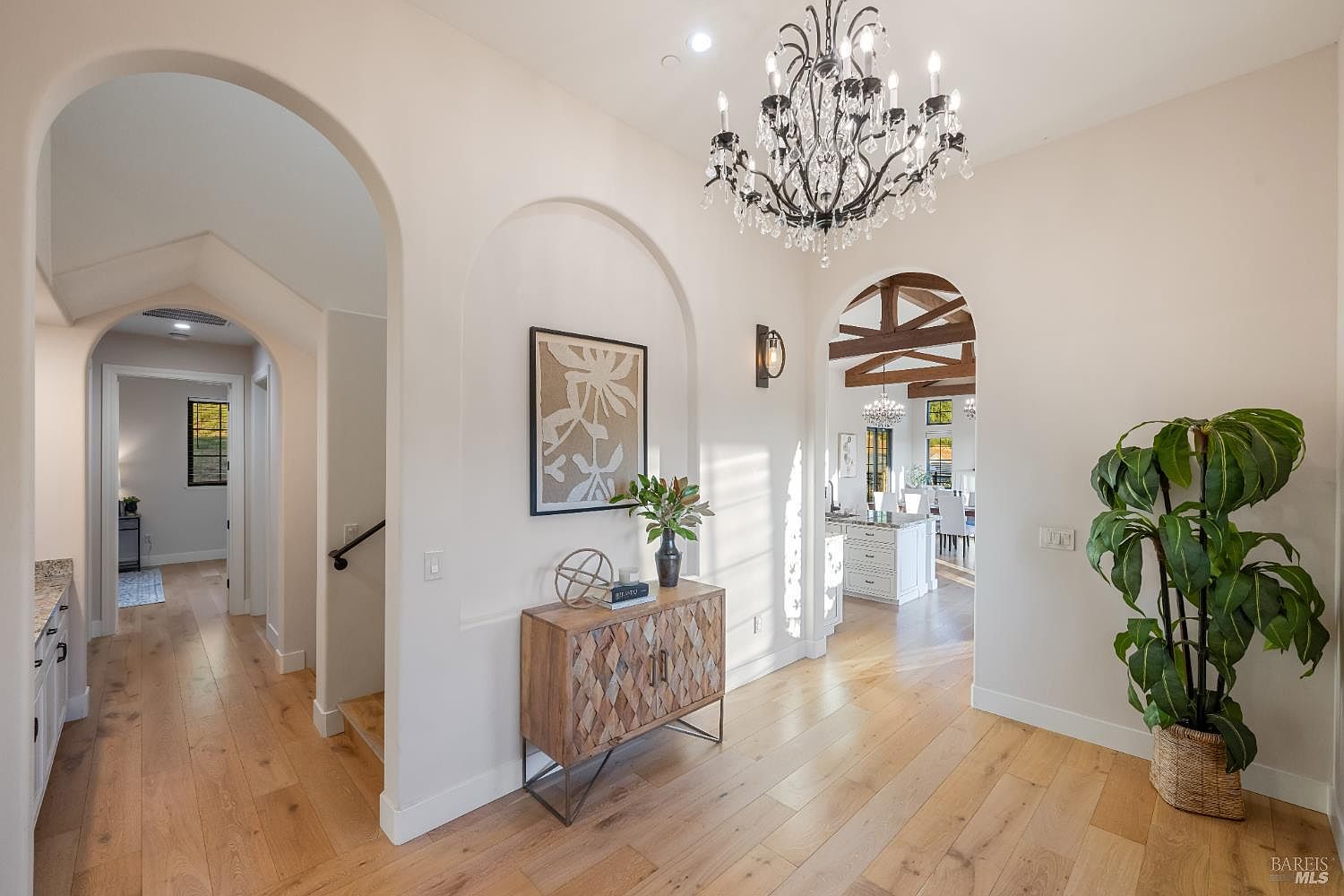 This interior shot showcases a well-lit hallway with hardwood floors and arched doorways, creating an elegant transition between spaces. A decorative cabinet with artwork and a large chandelier add sophistication, while a glimpse into the adjacent kitchen reveals a bright and open layout. The overall impression is one of refined taste and seamless flow.