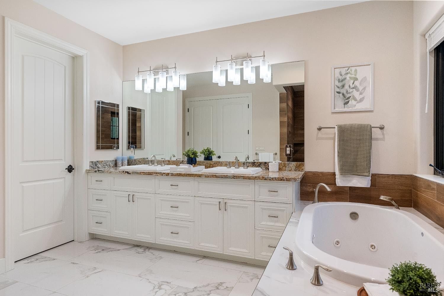 This is a primary bathroom featuring a double vanity with white cabinetry and a granite countertop. A large mirror spans the length of the vanity, illuminated by modern light fixtures. A soaking tub is visible in the foreground, accented by wood paneling and a marble surround, creating a luxurious and spa-like atmosphere.