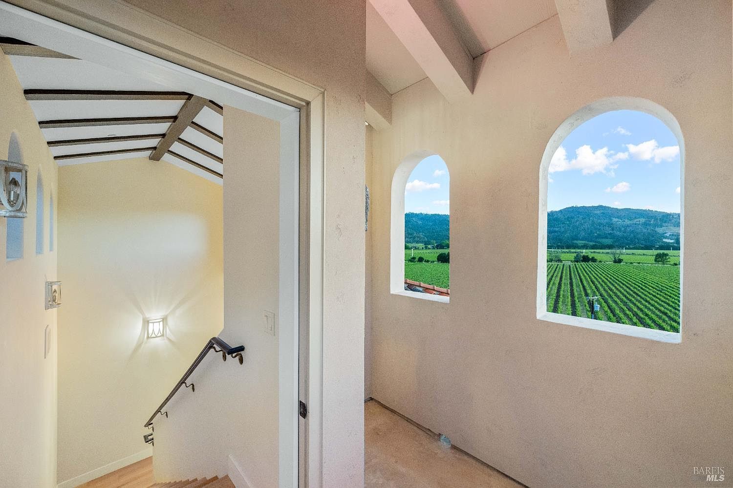 This interior shot showcases a hallway with a staircase leading upwards. The hallway features arched windows offering views of a vineyard landscape, while the staircase area is illuminated by wall sconces and natural light. The architectural details, including the ceiling beams and window design, add character to the space.