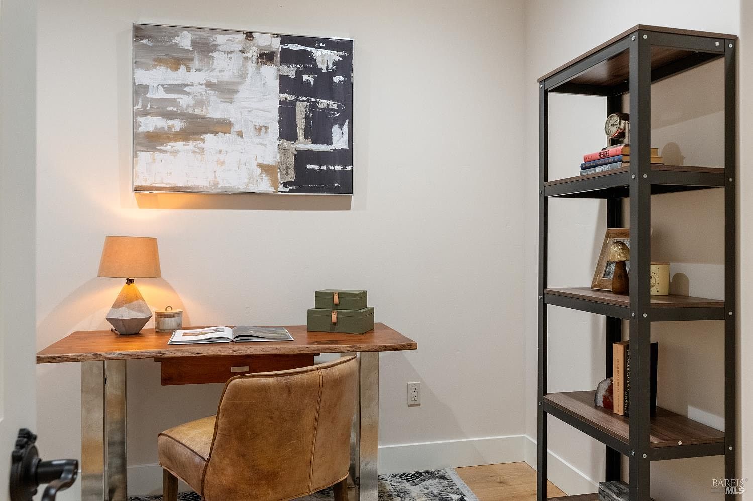 This is an interior shot of a home office or study area. The room features a wooden desk with metal legs, a leather chair, and a modern abstract painting on the wall. A tall, dark metal and wood bookshelf stands to the right, adding a touch of industrial chic to the space.