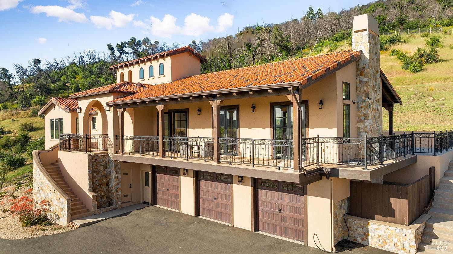 This is a front exterior view of a luxurious Mediterranean-style home. The house features a terracotta tile roof, stucco walls, and a multi-car garage. A wrought-iron balcony and stone accents add to the home's elegant appeal, while the surrounding landscape provides a sense of privacy and exclusivity.