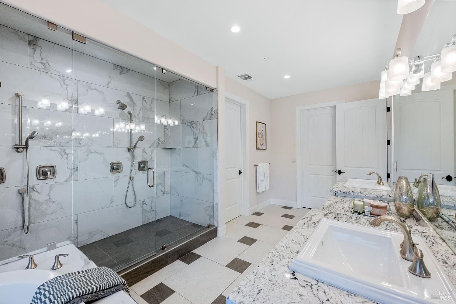 This is a well-lit primary bathroom featuring a glass-enclosed shower with marble tiling and dual shower heads. The bathroom includes a double vanity with white sinks and granite countertops, complemented by modern lighting fixtures. The flooring is a patterned tile, adding a touch of elegance to the space.
