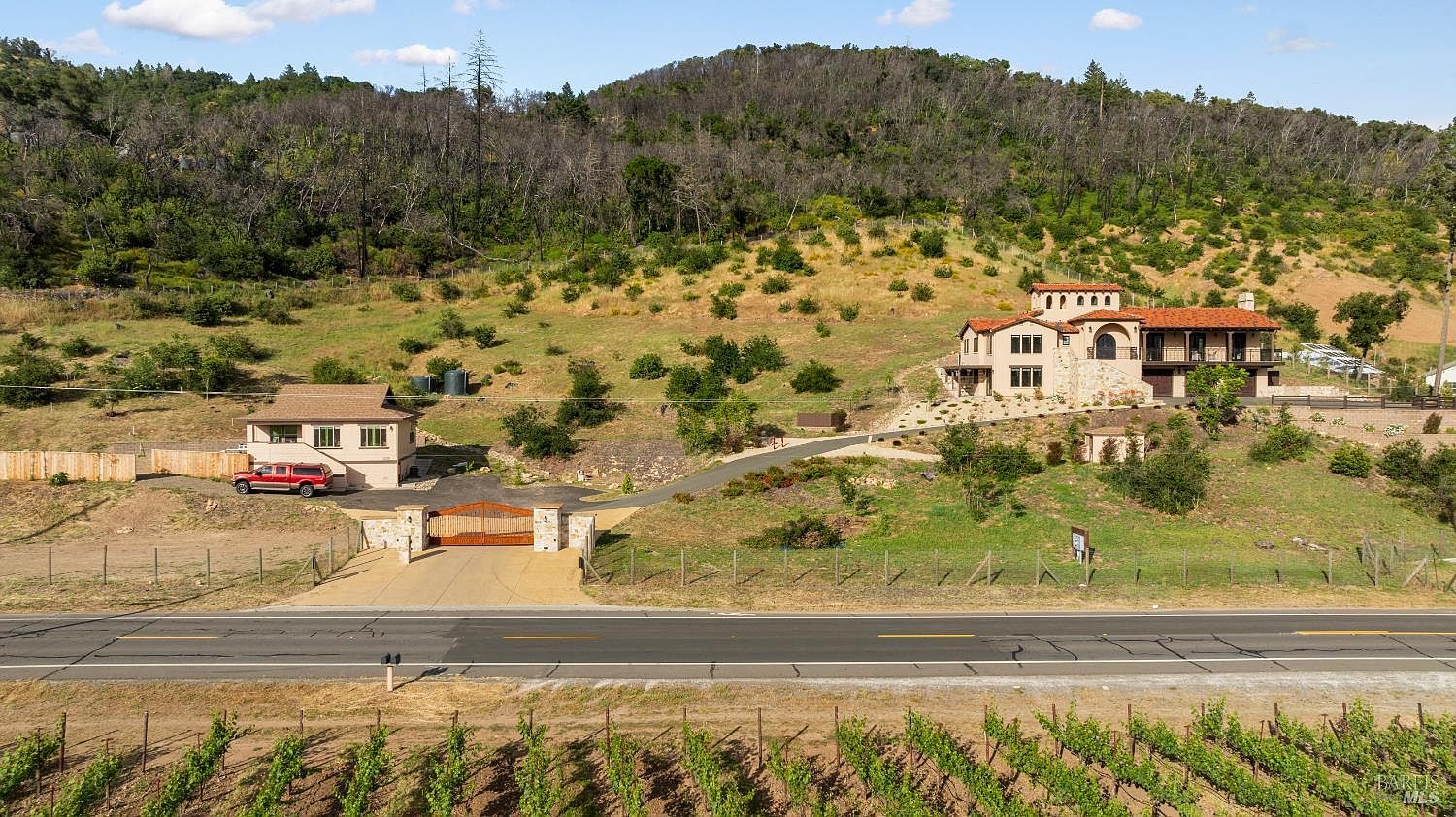 This aerial view showcases a luxurious estate featuring a Mediterranean-style main house with a red tile roof and stone accents, complemented by a separate guest house or auxiliary building. The property includes a gated entrance, a long driveway, and is set against a backdrop of rolling hills and vineyards, creating a sense of privacy and exclusivity. The perspective is cinematic, highlighting the scale and grandeur of the estate within its natural setting.