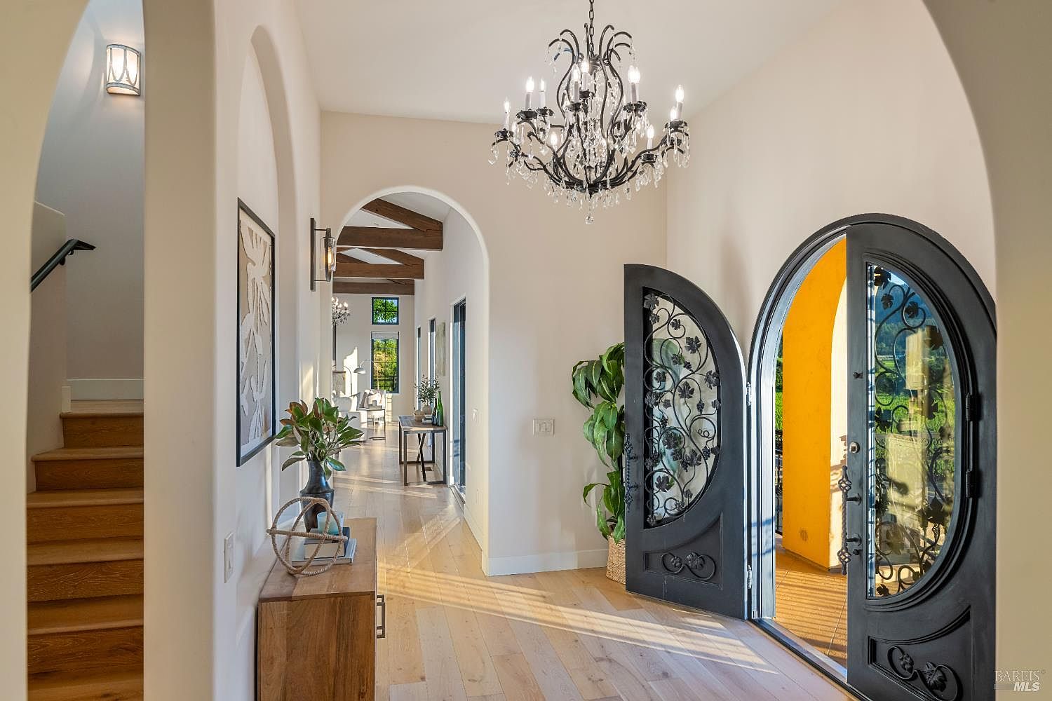 This interior shot showcases a grand hallway with arched doorways, a decorative chandelier, and an open front door with ornate ironwork. The hardwood floors and neutral wall colors create a bright and inviting space, while the staircase on the left adds architectural interest. The overall impression is one of elegance and spaciousness.