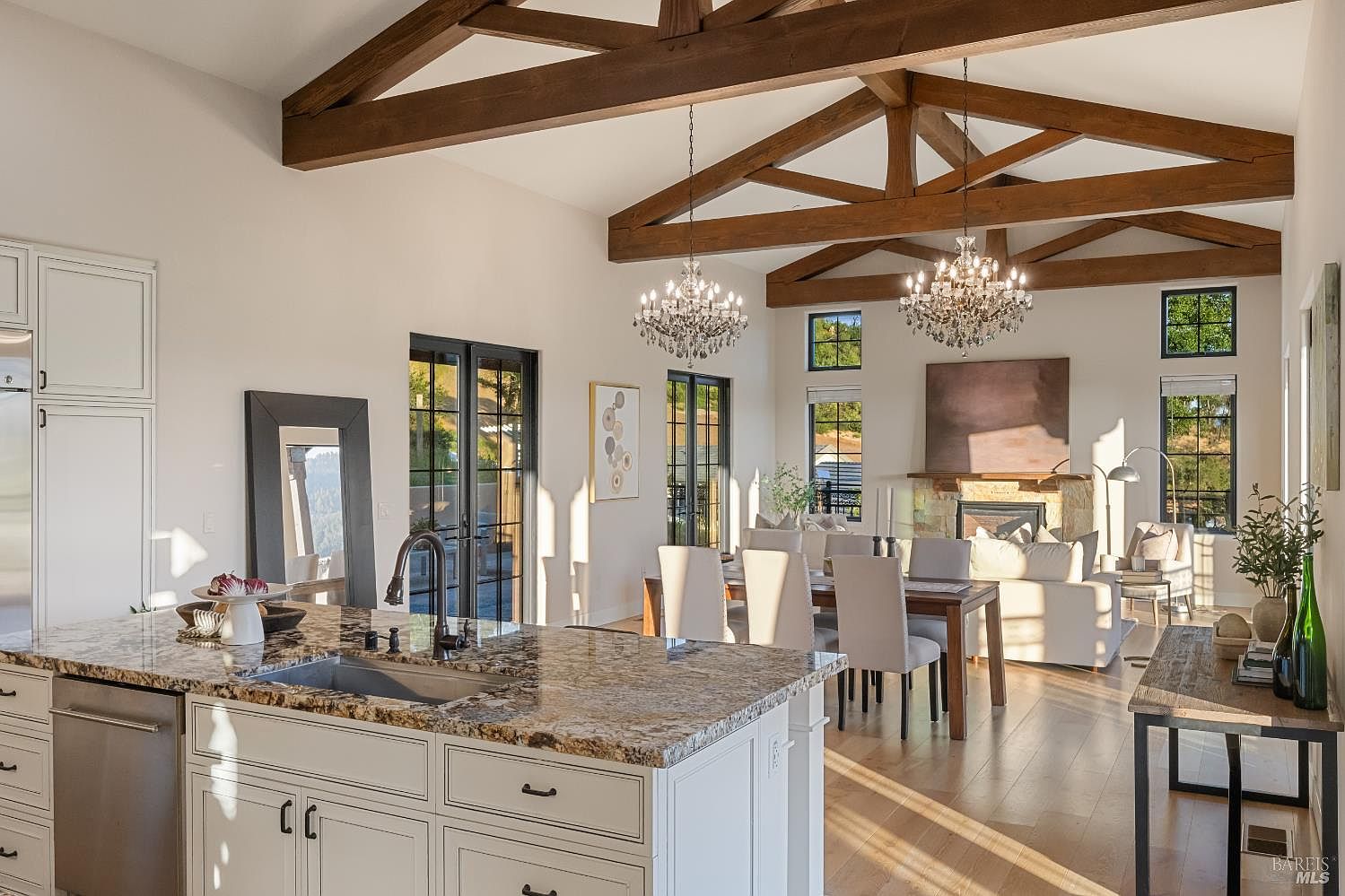 This interior shot showcases a bright and airy kitchen that seamlessly blends into the dining and living areas. The kitchen features a large island with granite countertops, white cabinetry, and stainless steel appliances. The open floor plan is accentuated by exposed wooden beams, elegant chandeliers, and large windows that flood the space with natural light, creating a warm and inviting atmosphere.