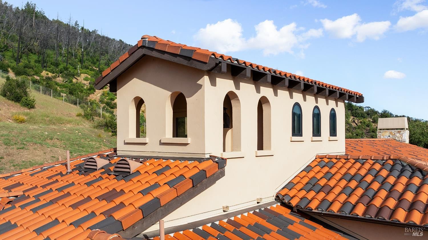 This image showcases the side of a Mediterranean-style building with a distinctive terracotta tile roof featuring alternating dark tiles. The building has arched windows and a light beige stucco exterior. The surrounding landscape includes trees and a glimpse of the sky, creating a serene and upscale impression.