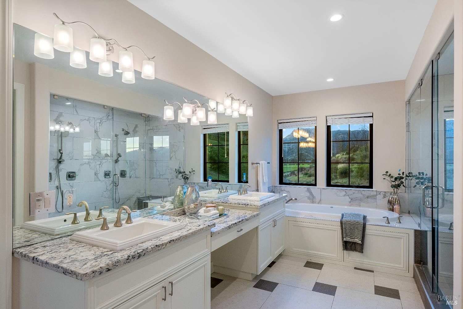 This is a luxurious primary bathroom featuring dual vanities with white cabinetry and granite countertops. A large mirror spans the length of the vanities, reflecting the natural light from the windows above the bathtub. The bathroom also includes a glass-enclosed shower and a freestanding tub, creating a spa-like atmosphere.