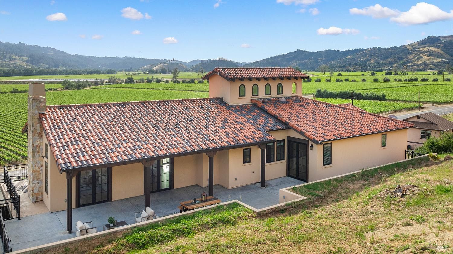 This aerial shot showcases a stunning single-story home with a terracotta tile roof, set amidst lush vineyards and rolling hills. The property features a covered patio with dark wood columns, a stone chimney, and a well-manicured lawn, creating a serene and luxurious estate. The house is surrounded by greenery, emphasizing its private and picturesque setting.