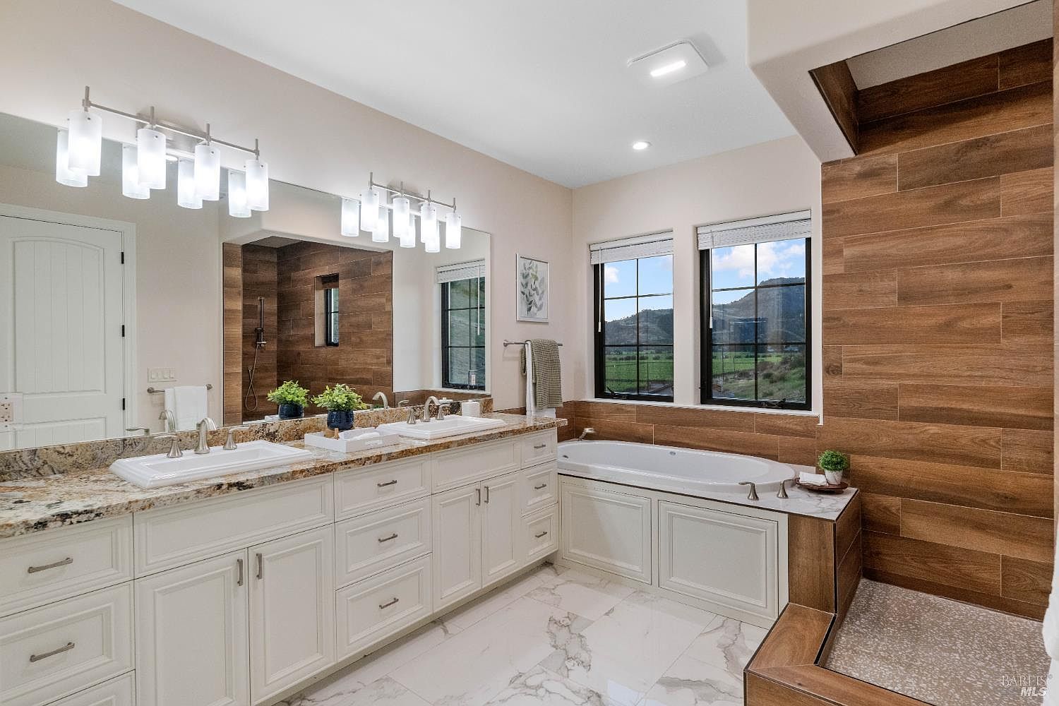 This is a primary bathroom featuring a double vanity with granite countertops and white cabinetry. A large mirror reflects the vanity and a walk-in shower with wood-look tile. A soaking tub is situated beneath two windows offering a view of the landscape, and the flooring is marble. The overall impression is luxurious and well-appointed.