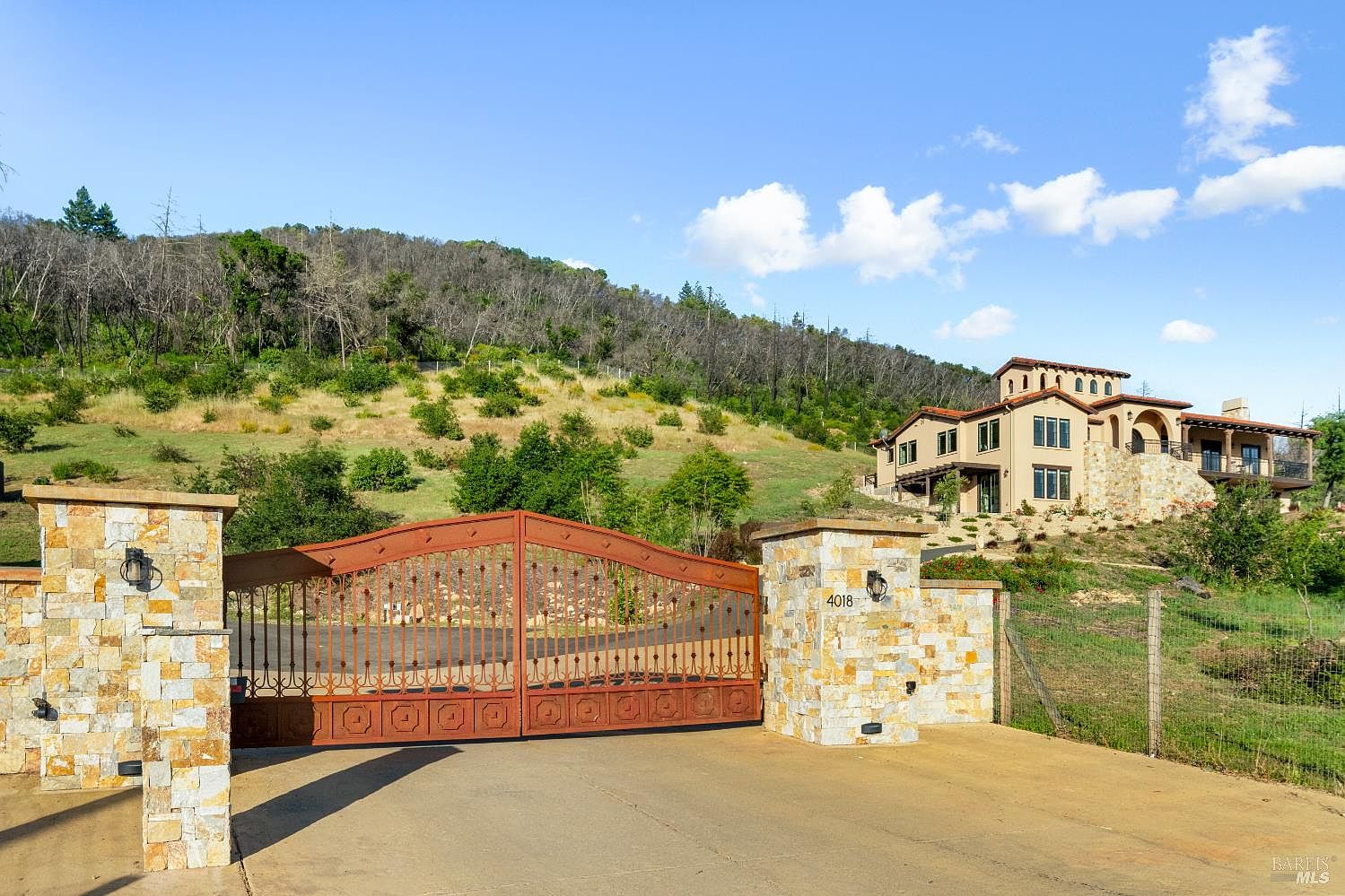 The image showcases the grand entryway of a property, featuring a stone-clad gatehouse with an ornate metal gate. The house is situated on a hill in the background, with a well-maintained landscape. The overall impression is one of security, privacy, and upscale living.