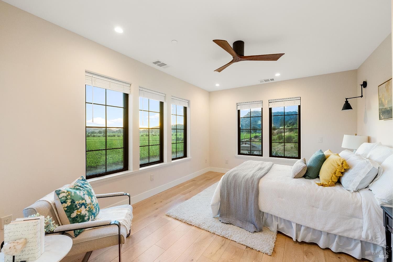 This is a bright and airy primary bedroom featuring hardwood floors, large windows with views of a green field, and a ceiling fan. The bed is neatly made with white linens and decorative pillows, and a cozy armchair sits in the corner. The room has a clean, modern aesthetic with neutral wall colors.