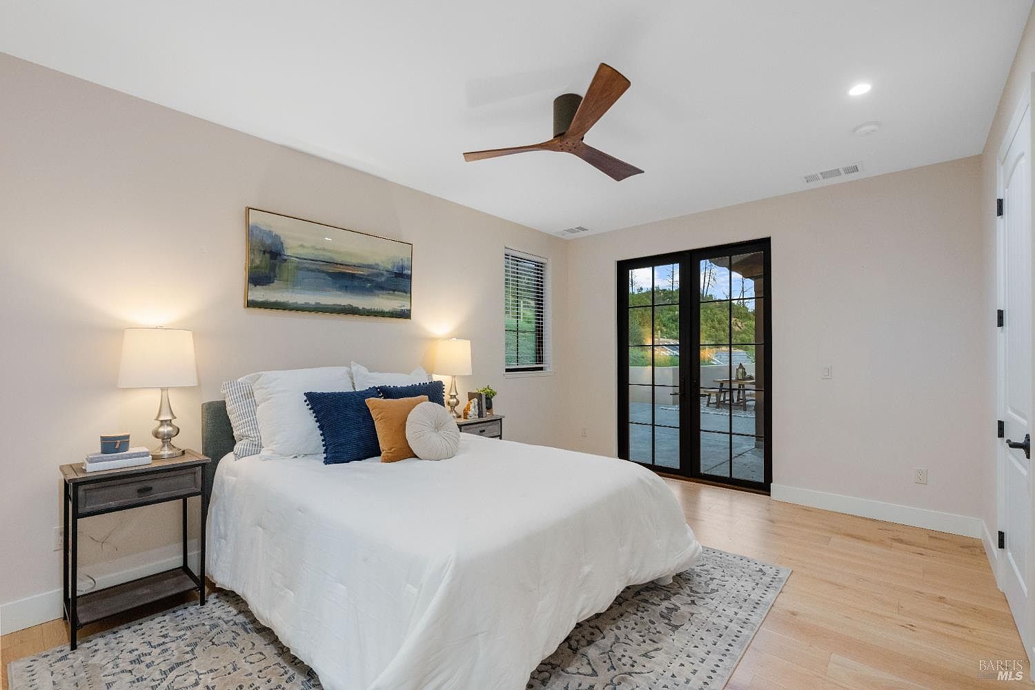 This is a bright and airy primary bedroom featuring a bed with white linens and decorative pillows, flanked by nightstands with lamps. A modern ceiling fan adds a touch of contemporary style, while a large glass door provides access to an outdoor area and fills the room with natural light. The hardwood floors and neutral wall color create a serene and inviting atmosphere.