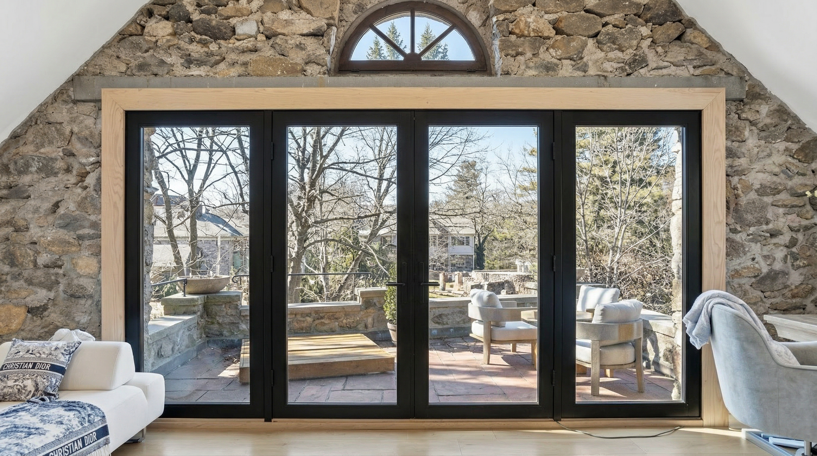 This living room features a rustic and refined design, highlighted by a stunning stone wall and a large, arched window. A massive four-panel black-framed door assembly provides direct access to a natural stone patio, seamlessly blending indoor and outdoor living. The space includes a comfortable sofa with patterned pillows and a light-filled, airy aesthetic that emphasizes the architectural character of the property.