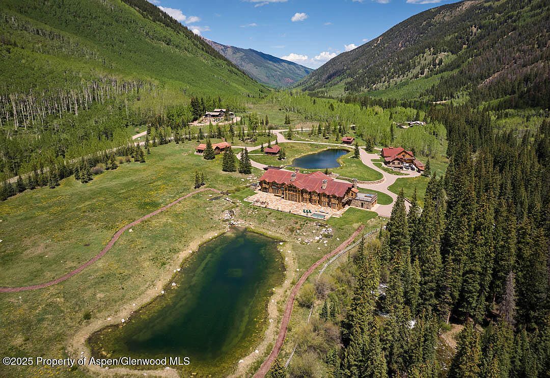 This aerial view showcases a sprawling estate nestled in a lush valley, surrounded by mountains and dense forests. The property features a large log-style home with a red roof, multiple outbuildings, and several ponds, all connected by winding pathways. The landscape is a mix of manicured lawns and natural terrain, creating a serene and private setting.