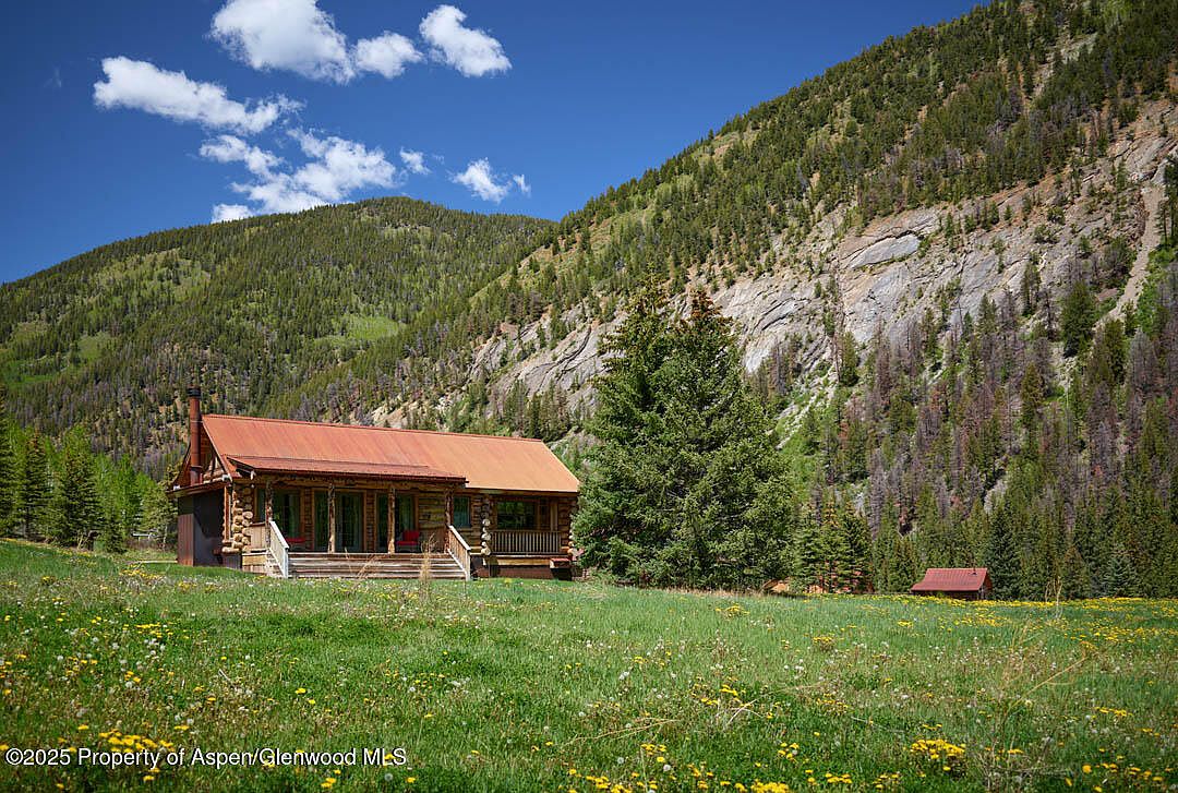 This image showcases a charming log cabin nestled in a picturesque mountain setting. The cabin features a rustic design with a red metal roof, a cozy porch, and a chimney. The surrounding landscape includes a lush green meadow dotted with wildflowers, towering evergreen trees, and a backdrop of rugged mountains, creating a serene and inviting atmosphere.