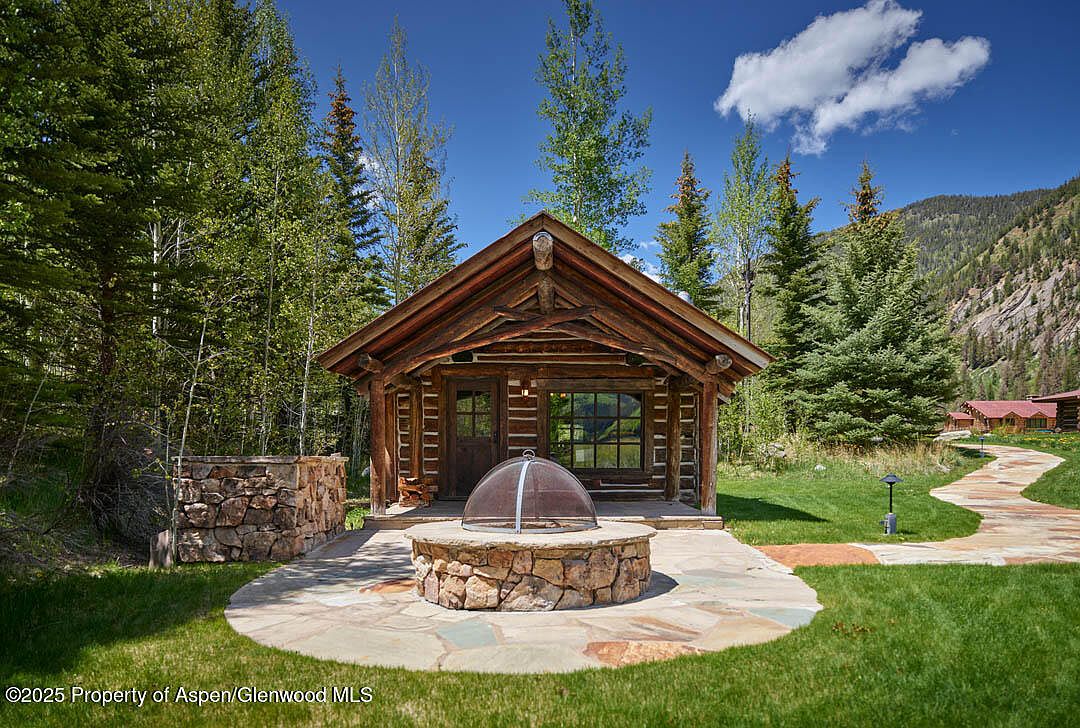 This image showcases the front exterior of a charming log cabin. The cabin features a rustic design with exposed wooden beams and a stone fire pit in the front yard. Lush greenery surrounds the cabin, creating a serene and inviting atmosphere, with a stone pathway leading to other buildings.