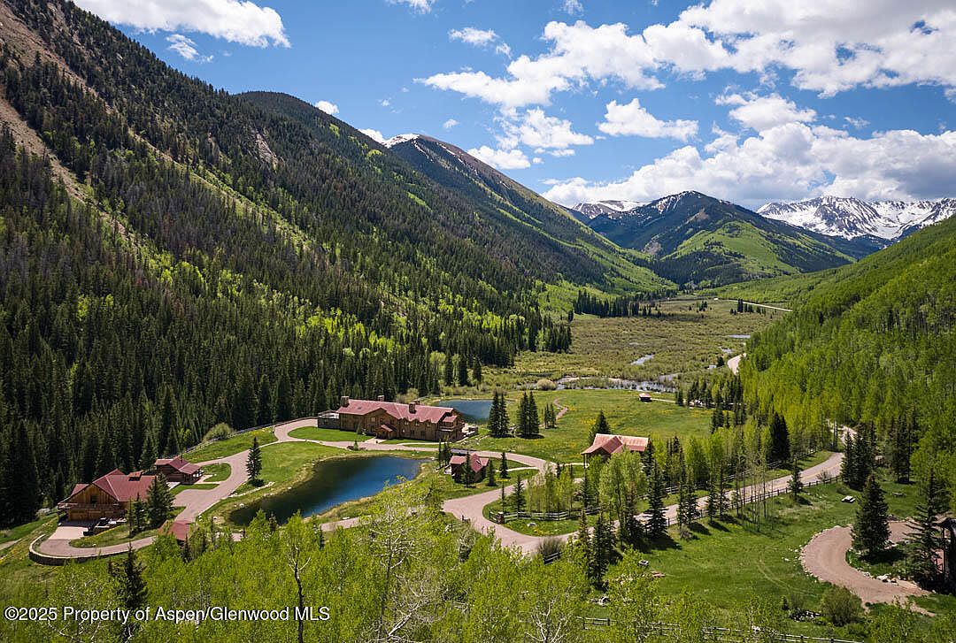 This aerial view showcases a luxurious estate nestled in a lush mountain valley. The property features multiple buildings with red roofs, surrounded by manicured lawns, ponds, and winding roads. The backdrop includes dense forests and snow-capped mountains, creating a serene and picturesque setting.