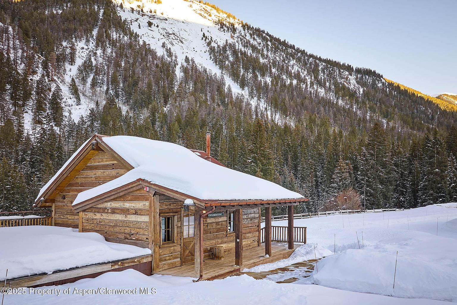 The image showcases a charming log cabin nestled in a snowy, mountainous landscape. The cabin features a covered porch, a snow-laden roof, and a rustic aesthetic that blends seamlessly with its natural surroundings. The scene evokes a sense of tranquility and seclusion, making it an ideal retreat.