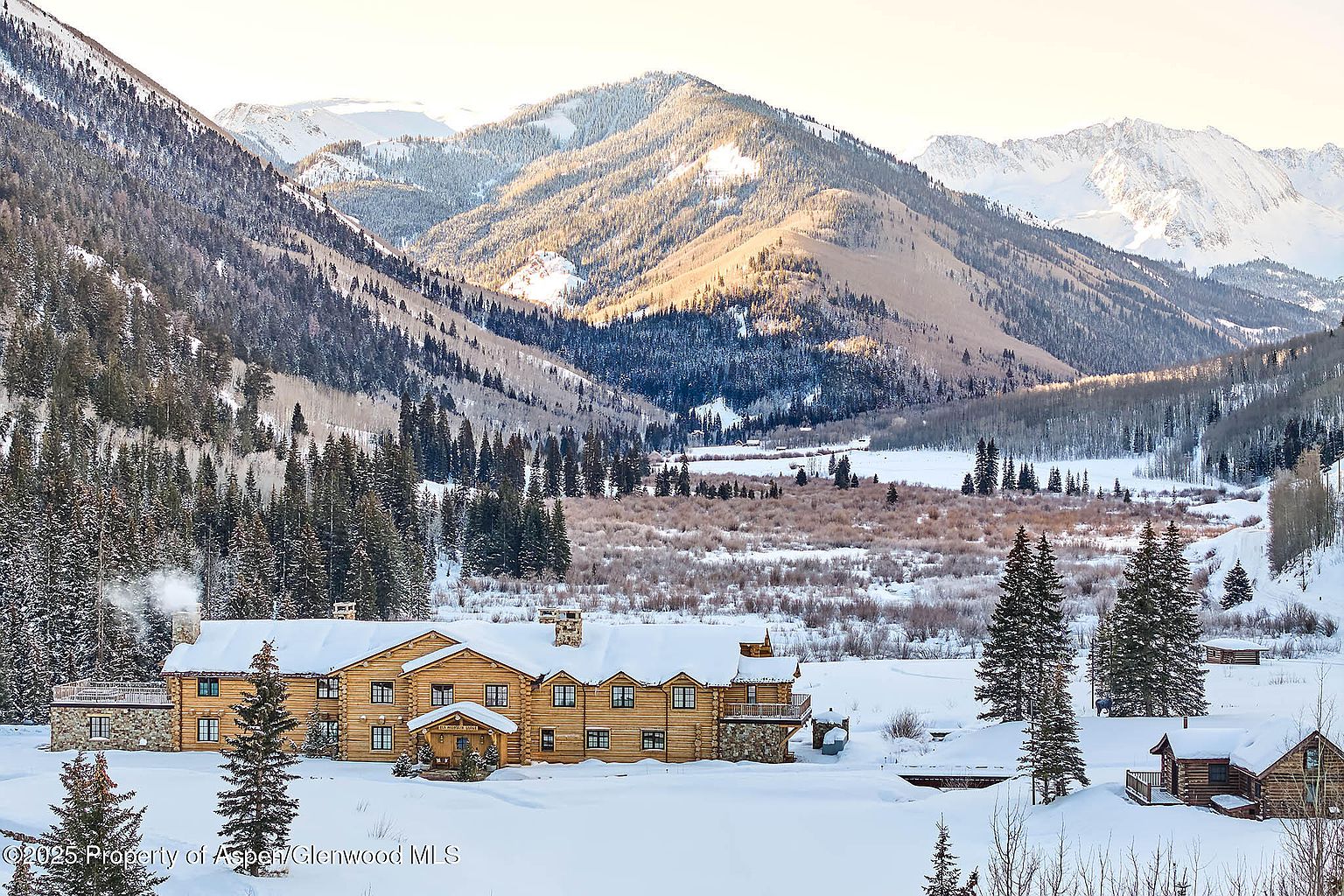 This image showcases the front exterior of a grand log cabin-style home nestled in a snowy mountain landscape. The home features multiple windows, stone accents, and a snow-covered roof, exuding a cozy and luxurious winter retreat. The surrounding snow-covered landscape and towering mountains create a picturesque and serene setting.