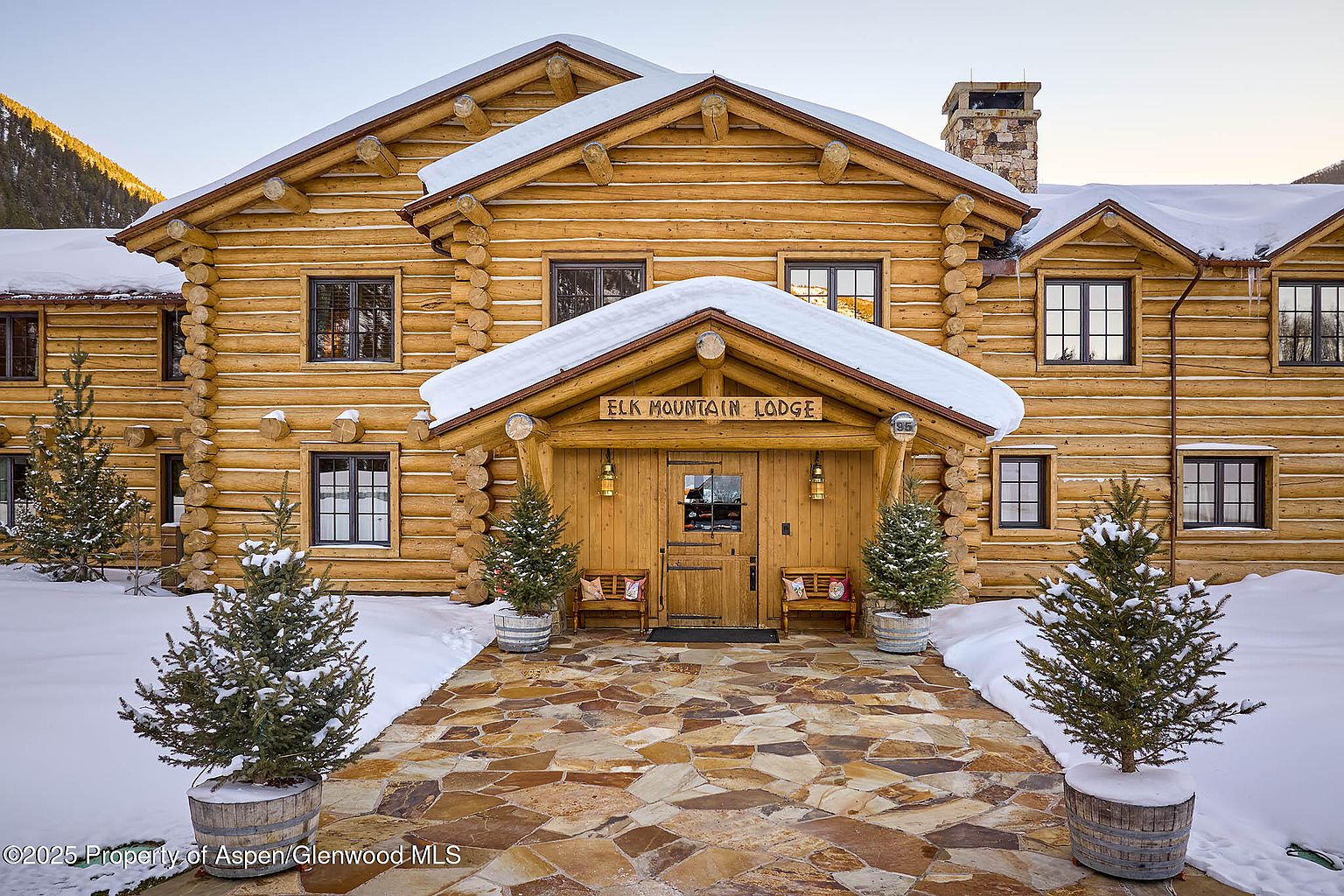 The image showcases the grand entryway of a log cabin-style lodge, featuring a stone pathway leading to a wooden door under a snow-covered porch with the sign "ELK MOUNTAIN LODGE". Flanking the entrance are small evergreen trees in wooden barrels, adding to the rustic charm and winter ambiance. The perspective is a medium shot, capturing the architectural details and inviting atmosphere.