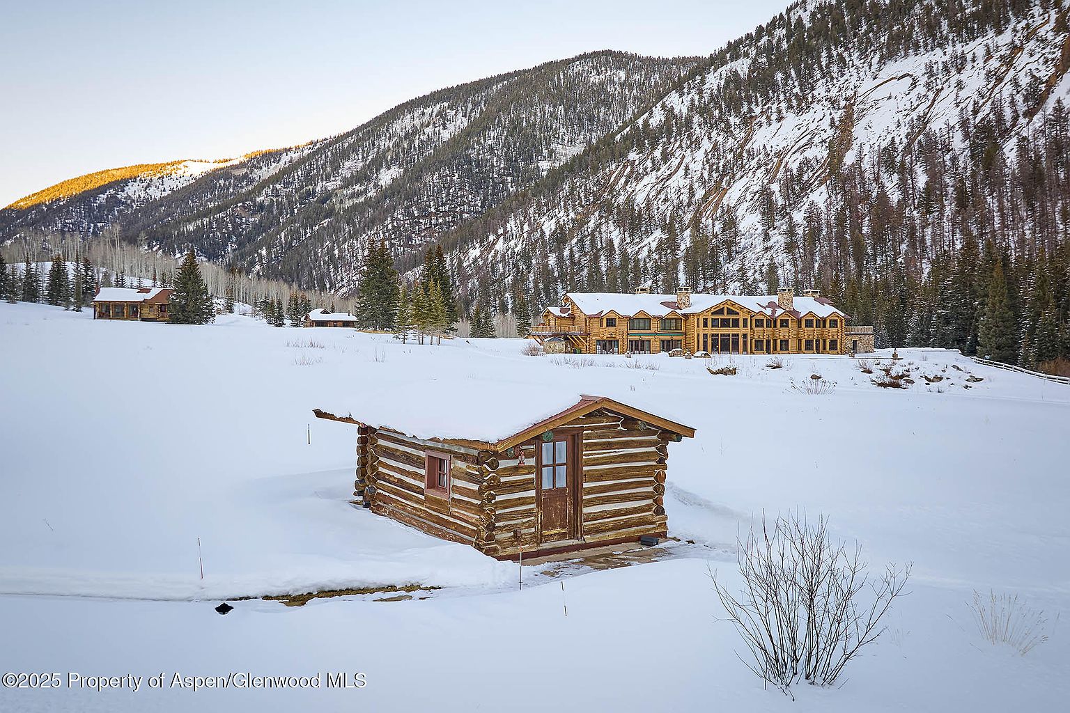 The image showcases a snow-covered landscape featuring a small log cabin in the foreground, with a larger log home visible in the background. The scene is set against a backdrop of a mountain partially covered in snow and trees. The overall impression is one of rustic charm and serene winter beauty, highlighting the potential for a secluded and picturesque property.