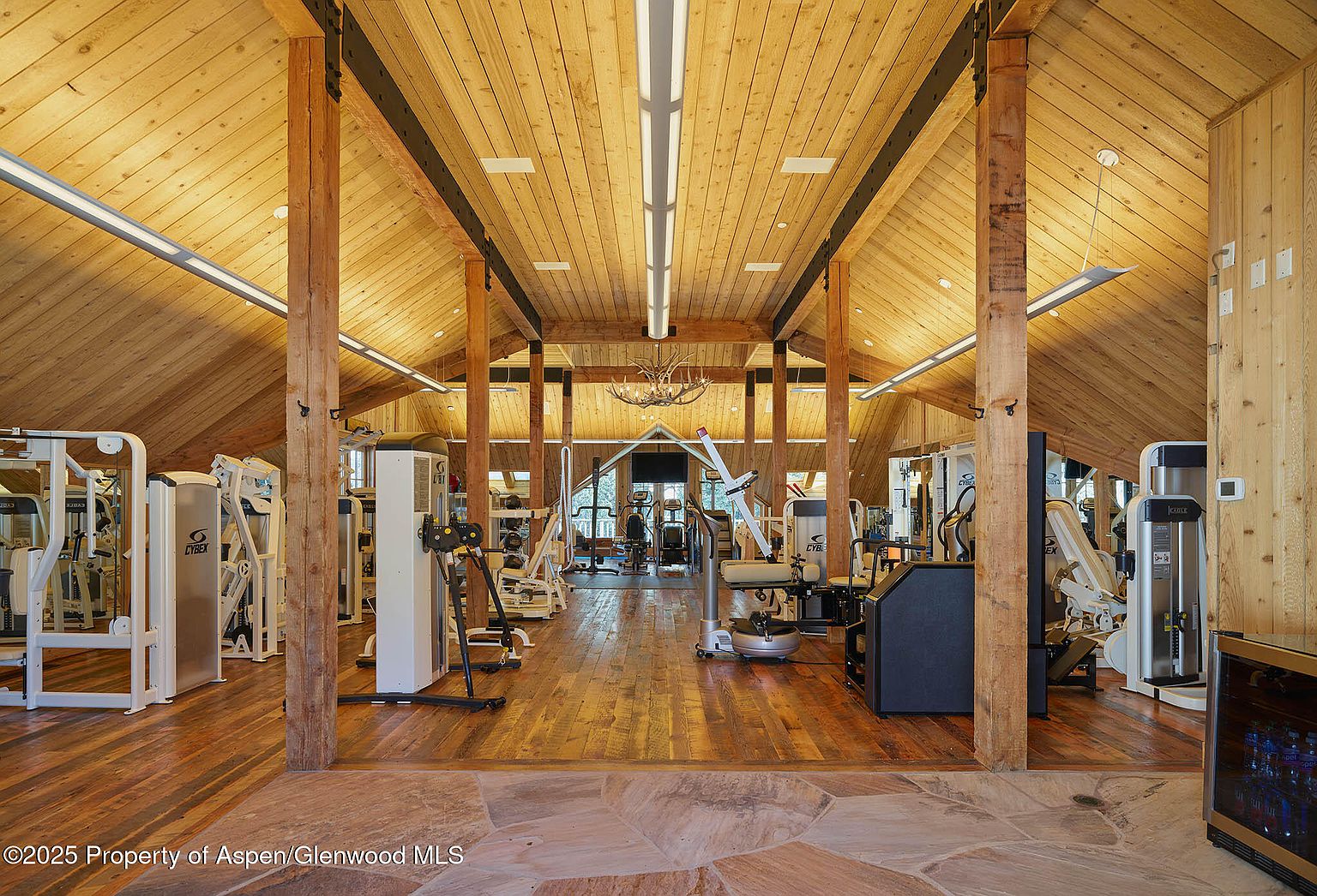 This is an interior shot of a spacious home gym featuring wood floors and a high, vaulted wood ceiling with exposed beams. Various Cybex weight machines are arranged throughout the room, and natural light filters in from a window at the far end. The overall impression is one of a well-equipped and inviting workout space.