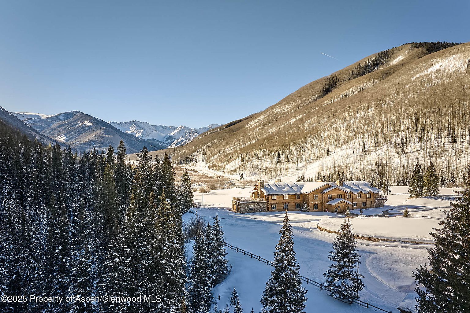 This aerial view showcases a luxurious log cabin nestled in a snowy mountain landscape. The cabin features a stone foundation, multiple chimneys, and a sprawling layout, blending seamlessly with the surrounding evergreen and aspen forests. The scene evokes a sense of secluded elegance and winter charm, ideal for a high-end real estate listing.