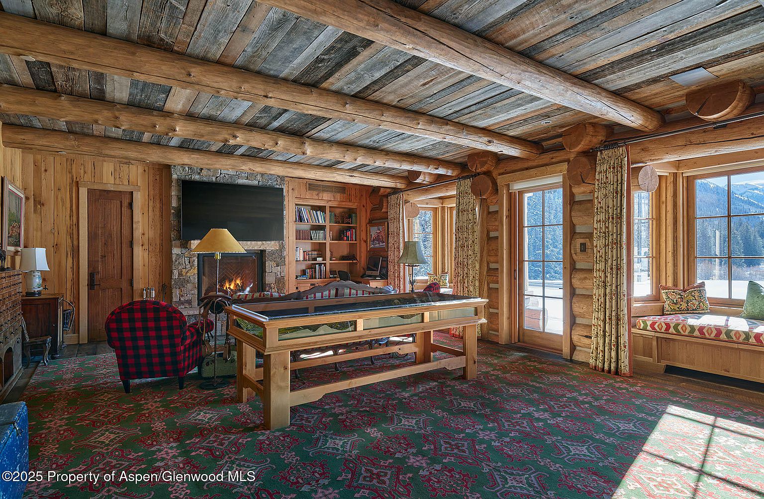 This is an interior shot of a rustic living room featuring log walls and ceiling beams. A stone fireplace with a television above it serves as a focal point, complemented by a plaid armchair and a unique display table. Natural light streams in through large windows, highlighting the room's warm and inviting atmosphere.