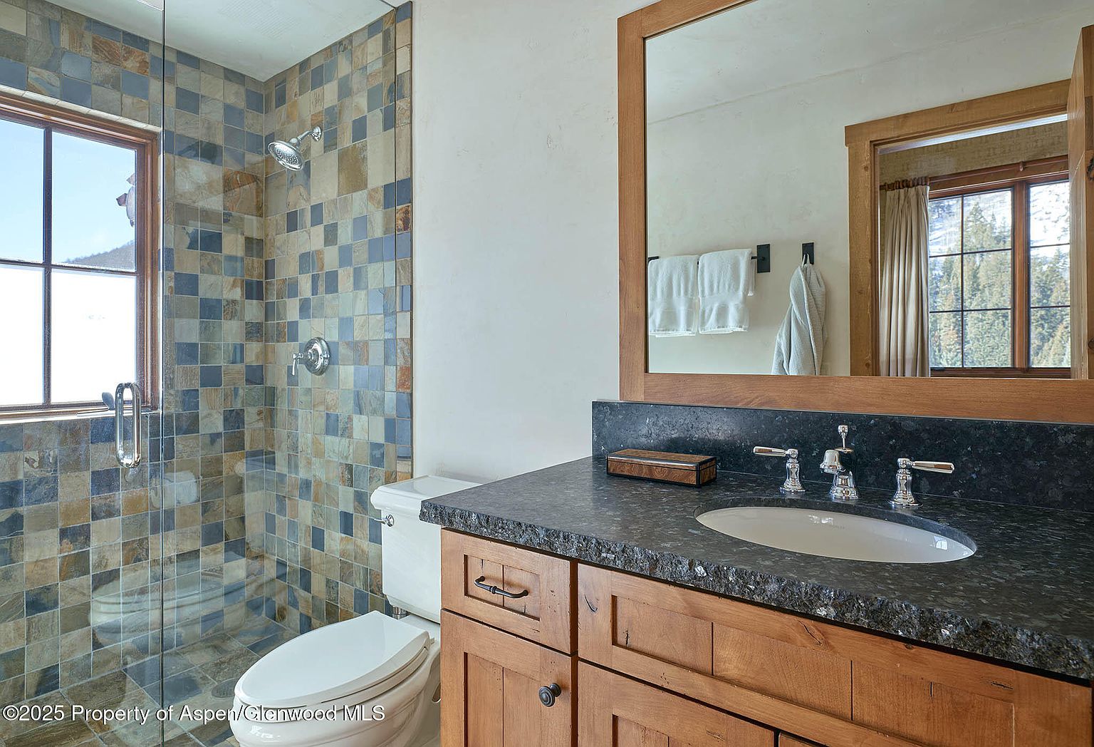 This is a bathroom featuring a tiled shower with a glass door, a toilet, and a wooden vanity with a dark countertop and a white sink. A framed mirror hangs above the vanity, reflecting a window and towels. The style is rustic and cozy, suitable for a mountain home.
